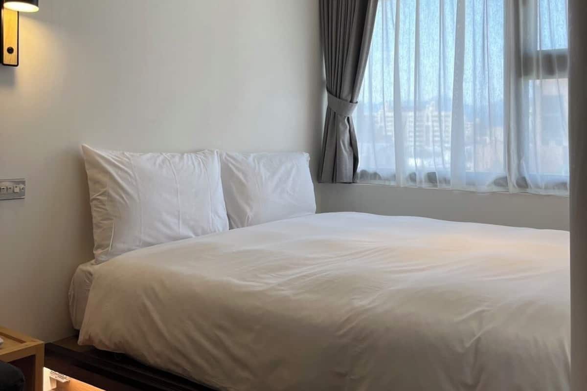 A hotel room with a large bed, a bedside table, a wall-mounted light, and a window with curtains.