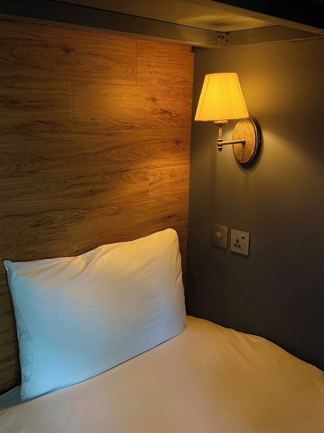 A hotel room with a wooden headboard, a white pillow, a wall-mounted lamp, and a power outlet.