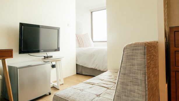 Una habitación pequeña y bien iluminada con una cama, un televisor sobre un mueble bajo, un refrigerador pequeño y una ventana que deja entrar luz natural.