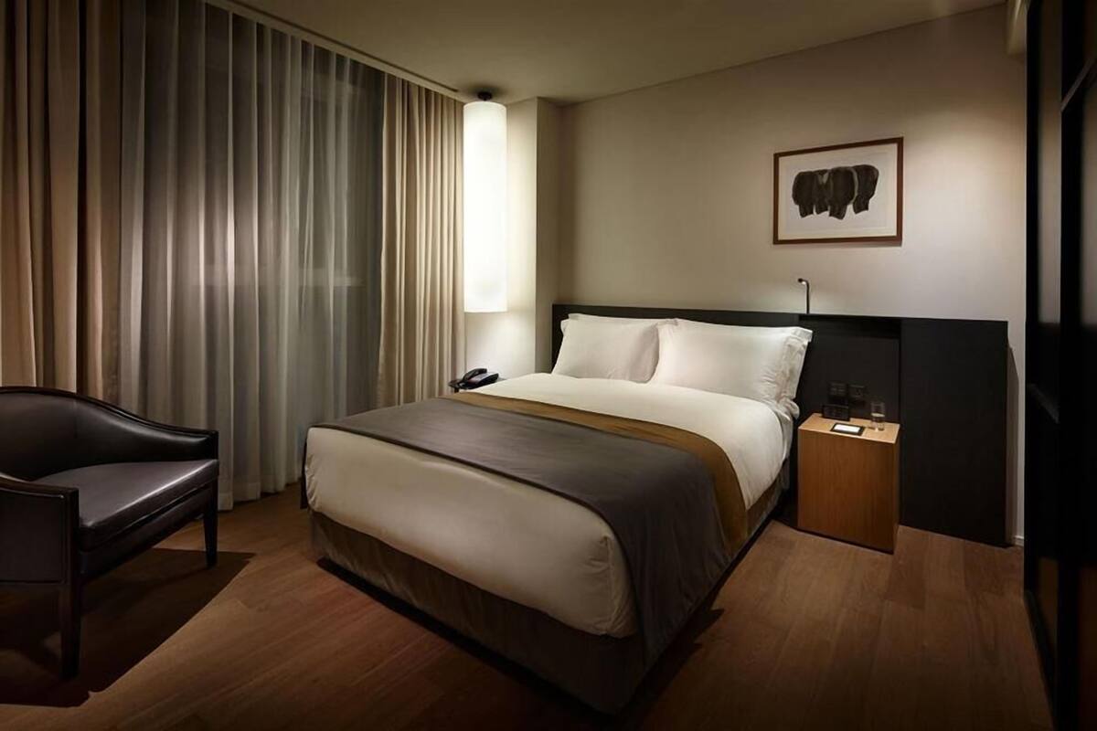 Una habitación de hotel con cama, una silla, mesita de noche con lámpara, teléfono y un cuadro en la pared.
