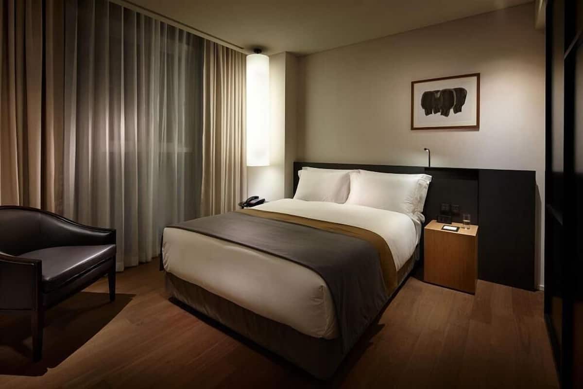 A hotel room with a bed, a chair, a bedside table with a lamp, a phone, and a framed picture on the wall.