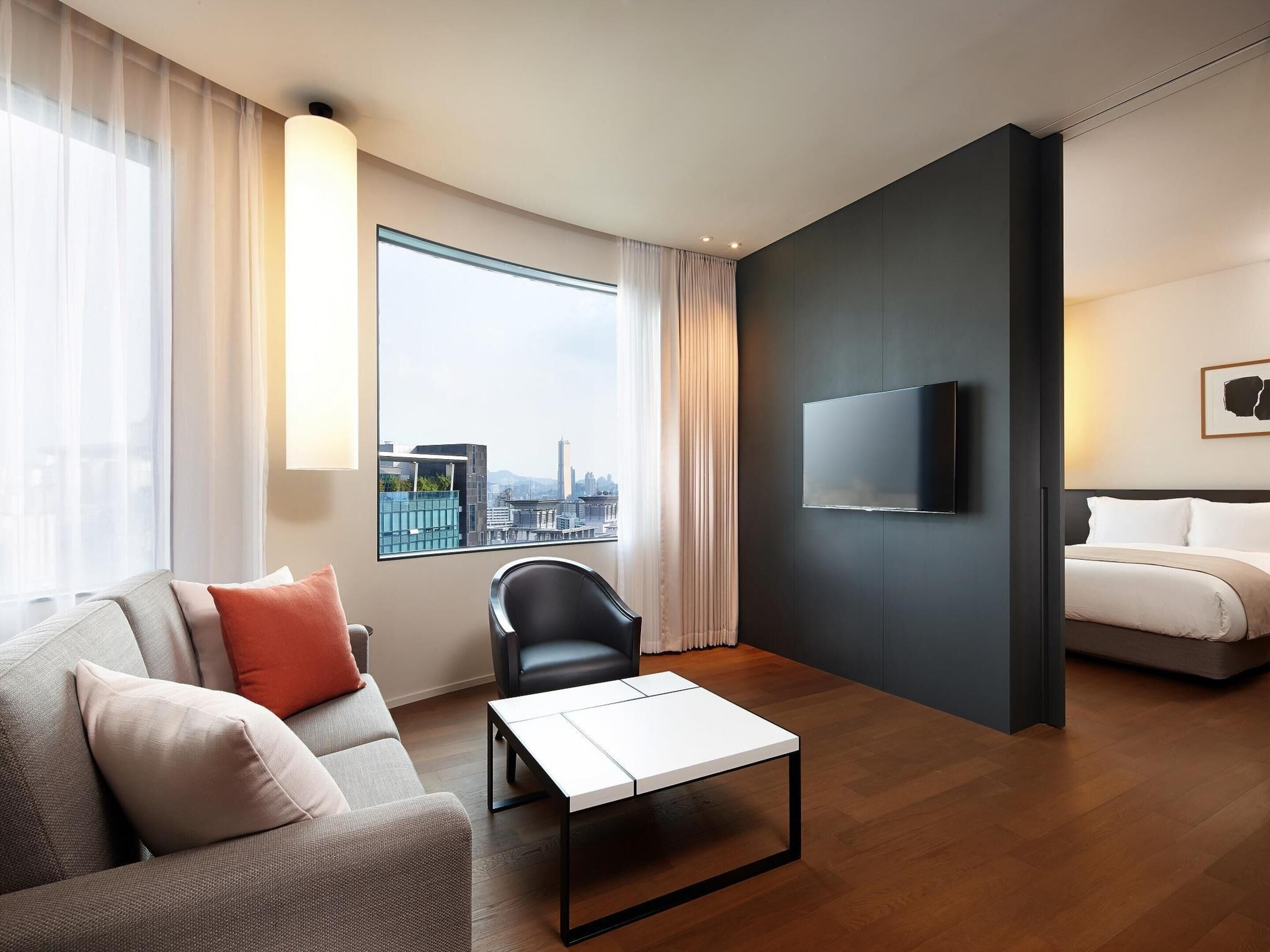A modern hotel room with a sofa, a chair, a coffee table, a bed, a TV, and a large window with a city view.