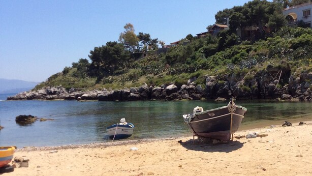 A sandy beach with two boats, one anchored and one on the shore, near a rocky shoreline with trees and a building in the background. - Case Corona (Santa Flavia)