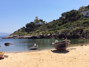 A sandy beach with two boats, one anchored and one on the shore, near a rocky shoreline with trees and a building in the background. - Case Corona (Santa Flavia)