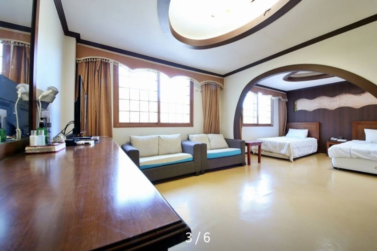 Een hotelkamer met een houten bureau, een bank, twee enkele bedden en een groot raam met gordijnen.