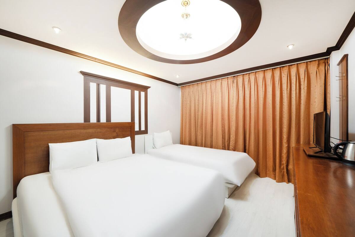 Een hotelkamer met twee bedden, een houten hoofdbord, een groot raam met luxaflexen, een plafondlamp en een televisie op een houten tafeltje.