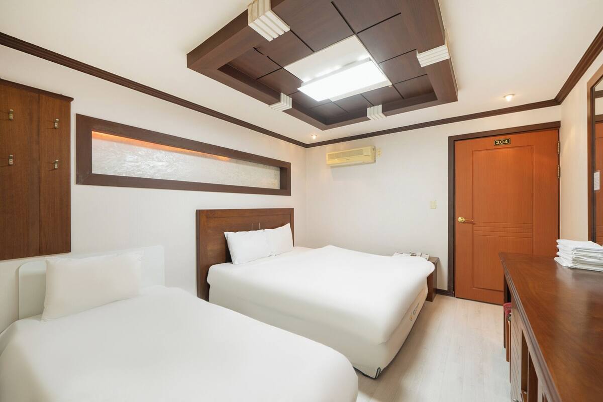 Hotelkamer met twee bedden, een houten hoofdbord, een plafond met inbouwspots, een airconditioning en een houten deur met een nummer.