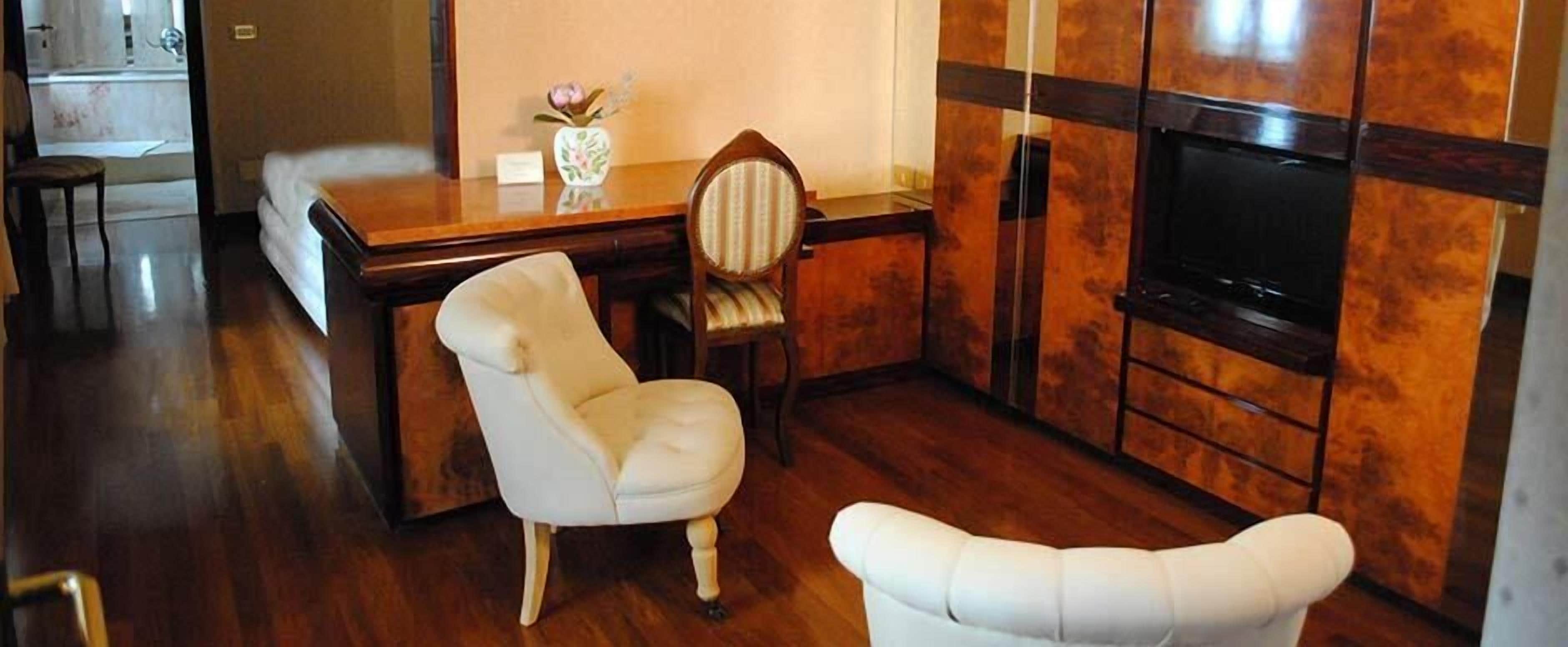A room with a wooden floor, a white armchair, a wooden desk with a vase of flowers, and a television mounted on the wall.