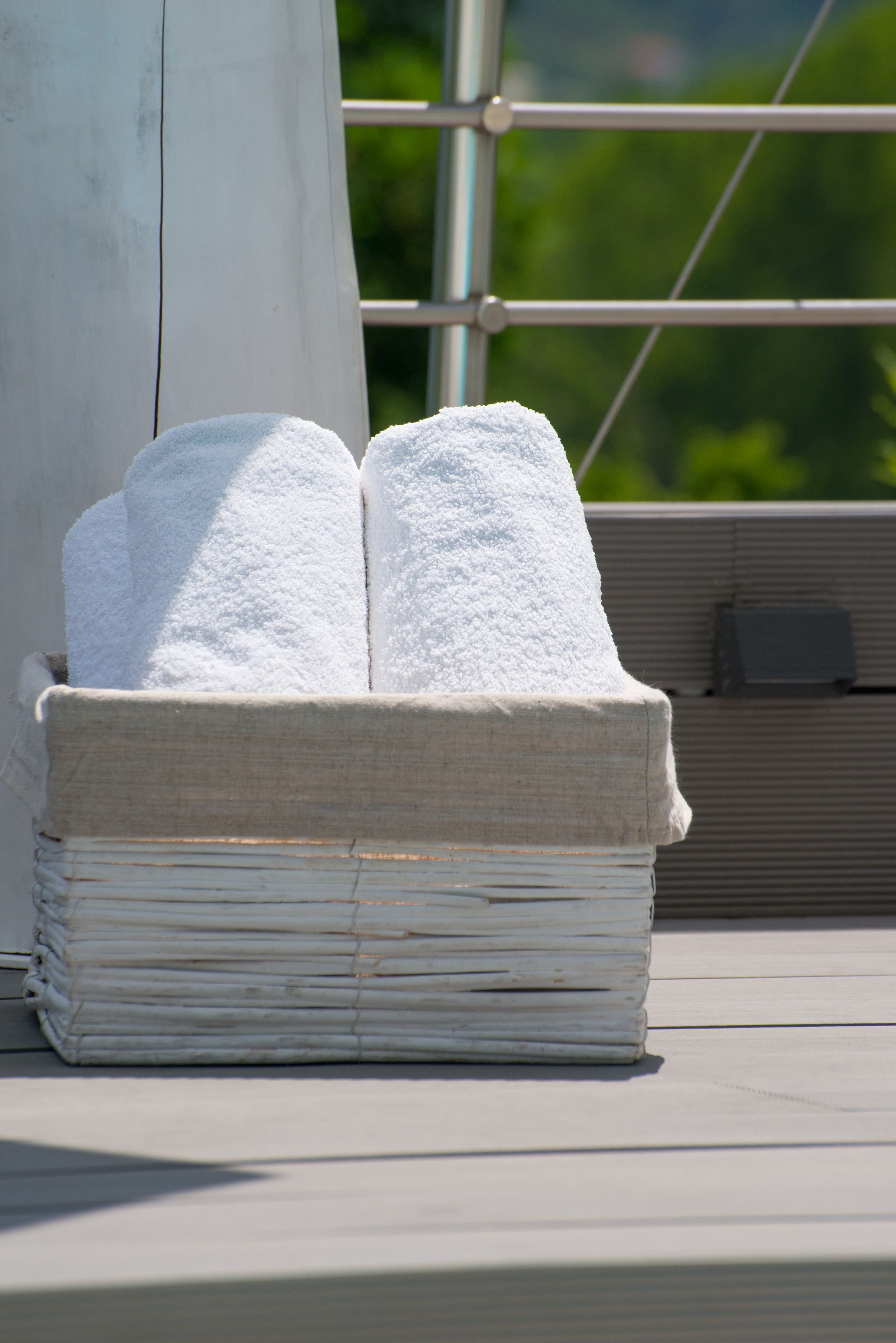A white, rectangular, woven basket with two white towels stacked on top, placed on a balcony with a metal railing and greenery in the background.