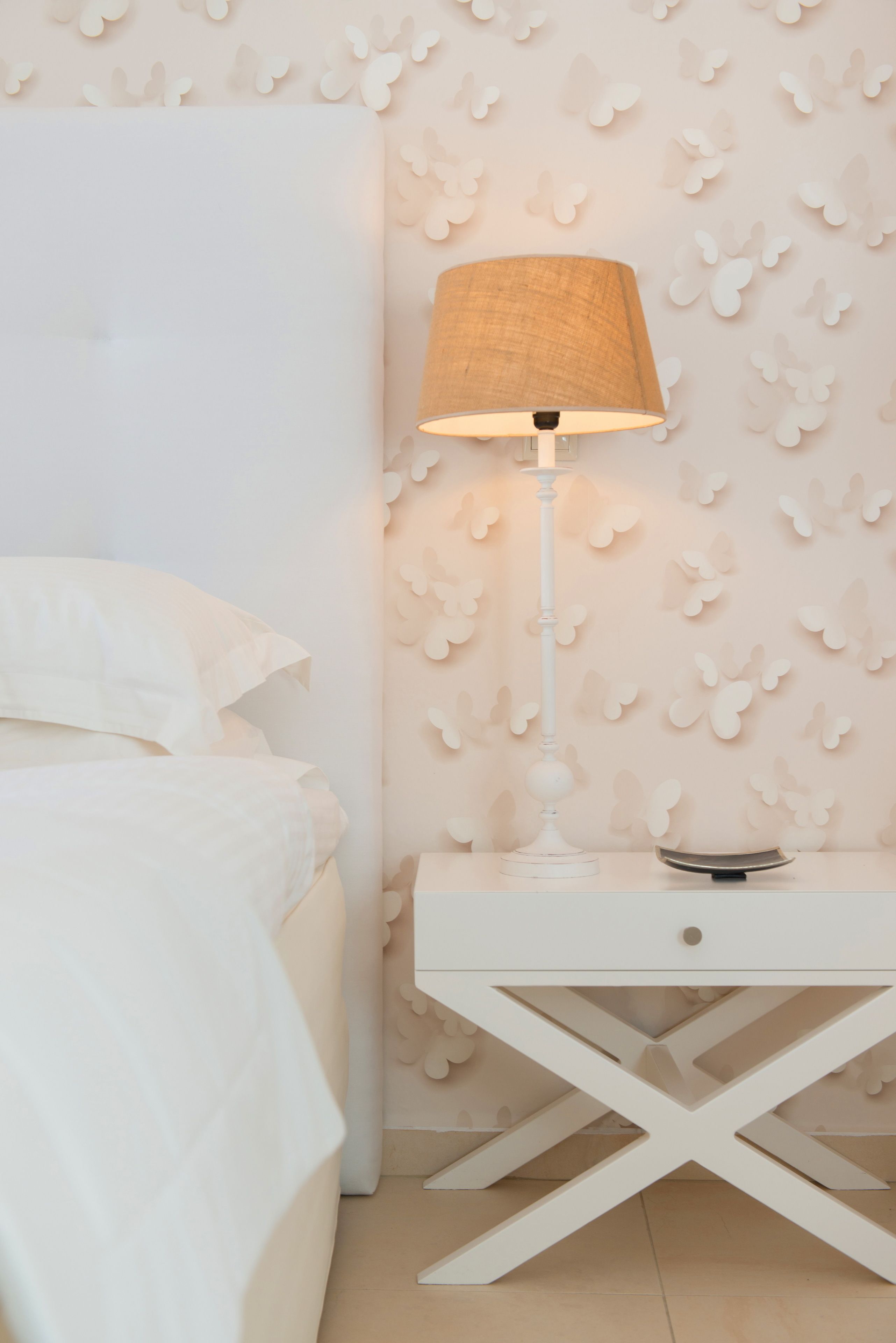 Una cama bien hecha con ropa de cama blanca, una mesita de noche blanca con una lámpara, y una pared con un diseño de mariposas.