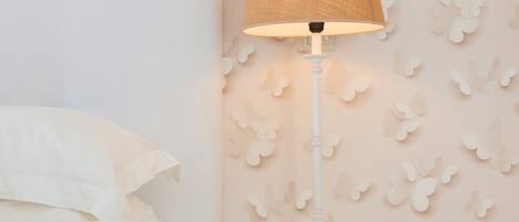 Una cama bien hecha con ropa de cama blanca, una mesita de noche blanca con una lámpara, y una pared con un diseño de mariposas.