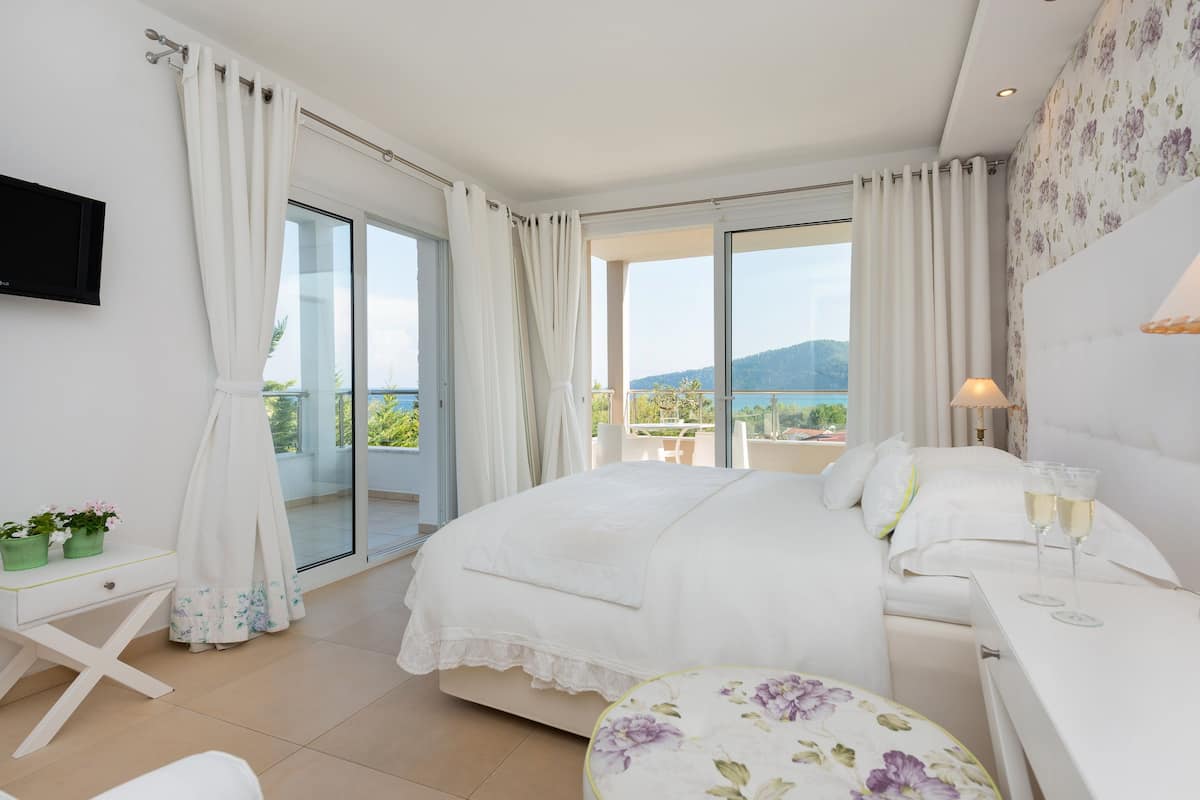 A bedroom with a large bed, a desk, a chair, a TV, and a balcony with a view of the mountains.