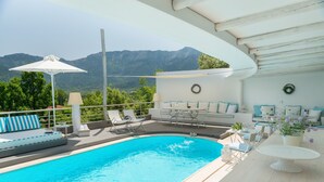 Un área de piscina con vistas a montañas, un espacio de descanso con sillas y una mesa, y un techo con vigas de madera.