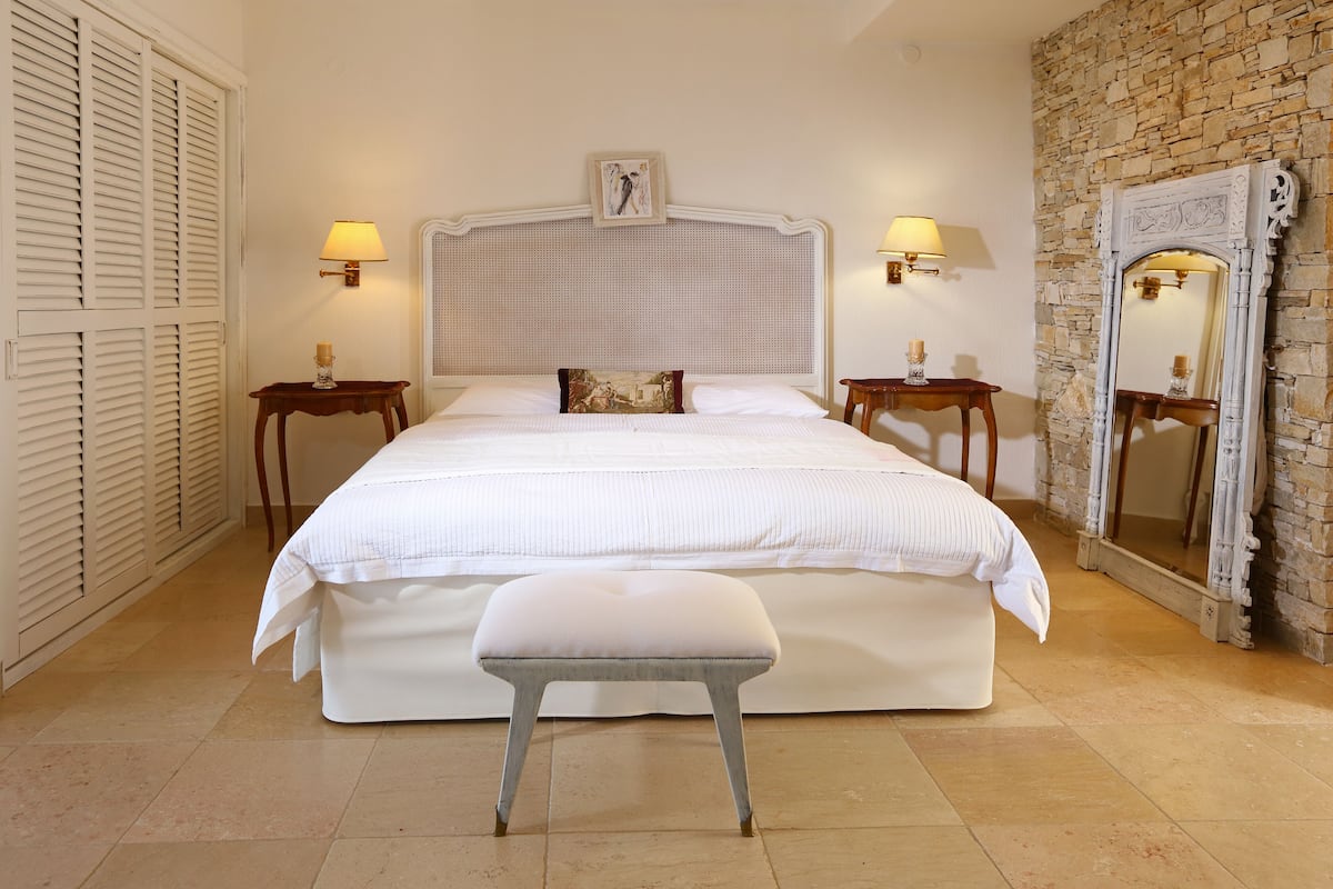 A bedroom with a large bed, two bedside tables with lamps, a bench, a wall-mounted picture, and a stone wall with a mirror.