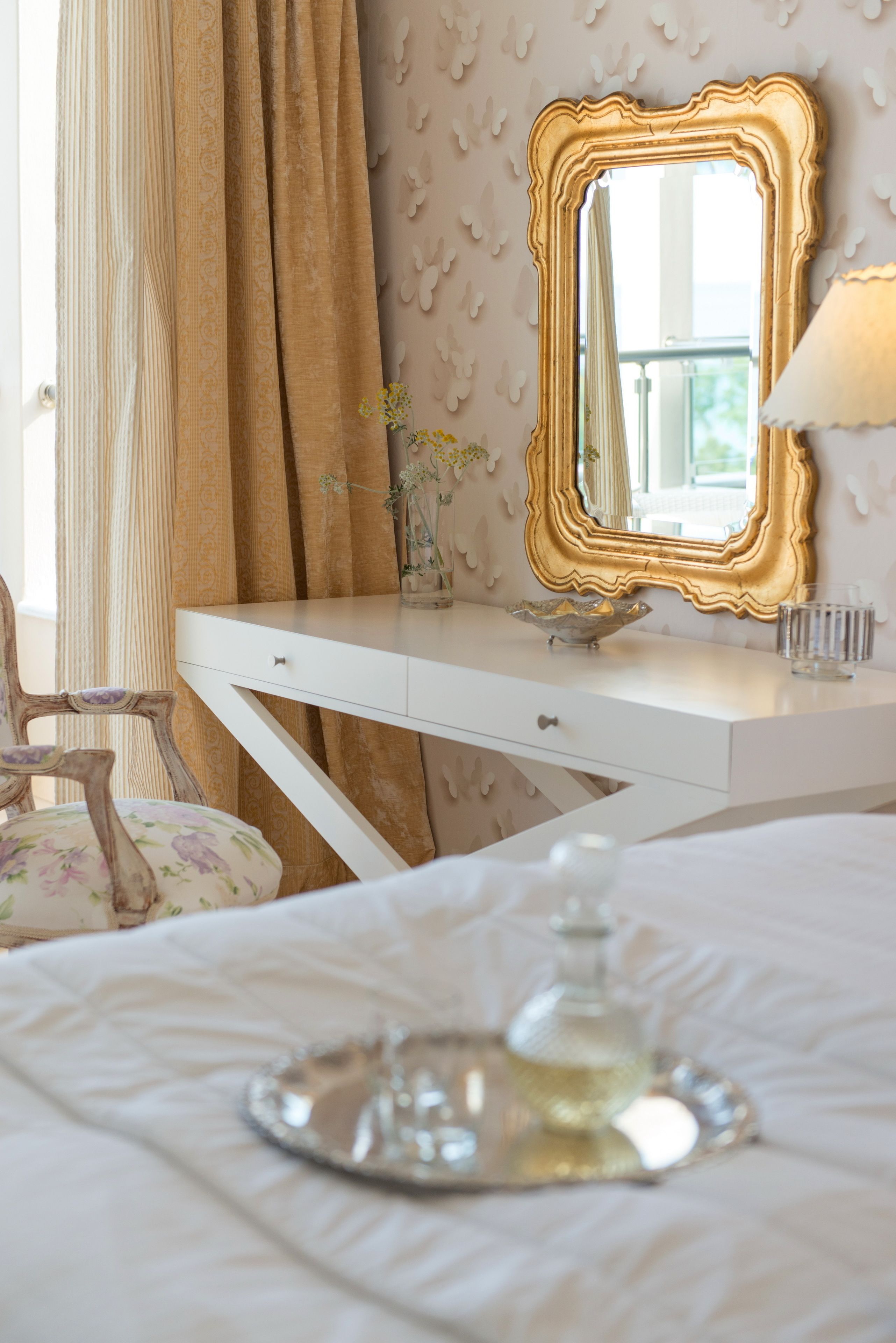 Een slaapkamer met een witte kaptafel, een spiegel met gouden lijst, een stoel met bloemenmotief en een nachtkastje met een glazen karaf.