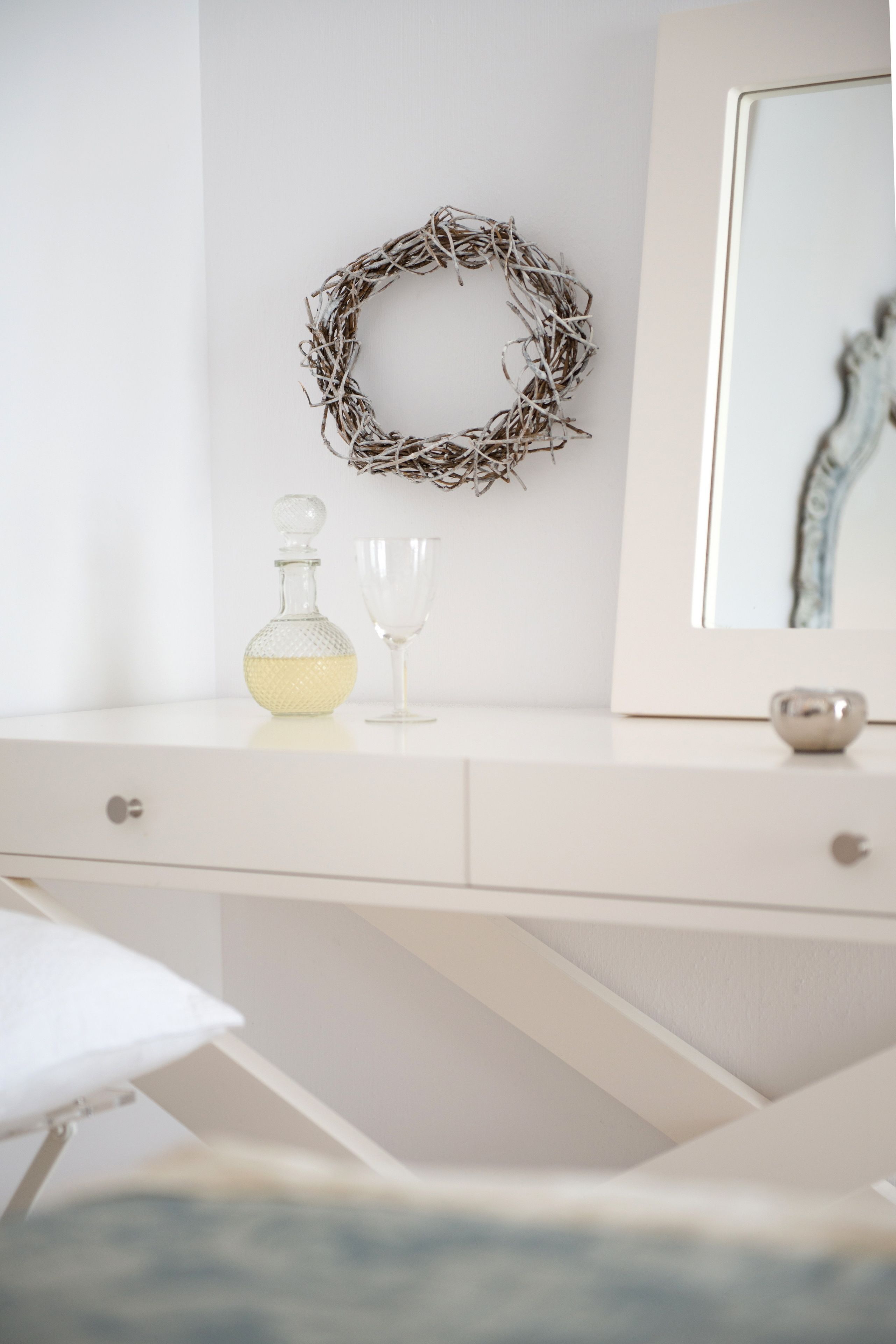 Une commode blanche sur laquelle sont posés un pichet en verre et un verre à vin. Une couronne tressée est accrochée au mur au-dessus de la commode.