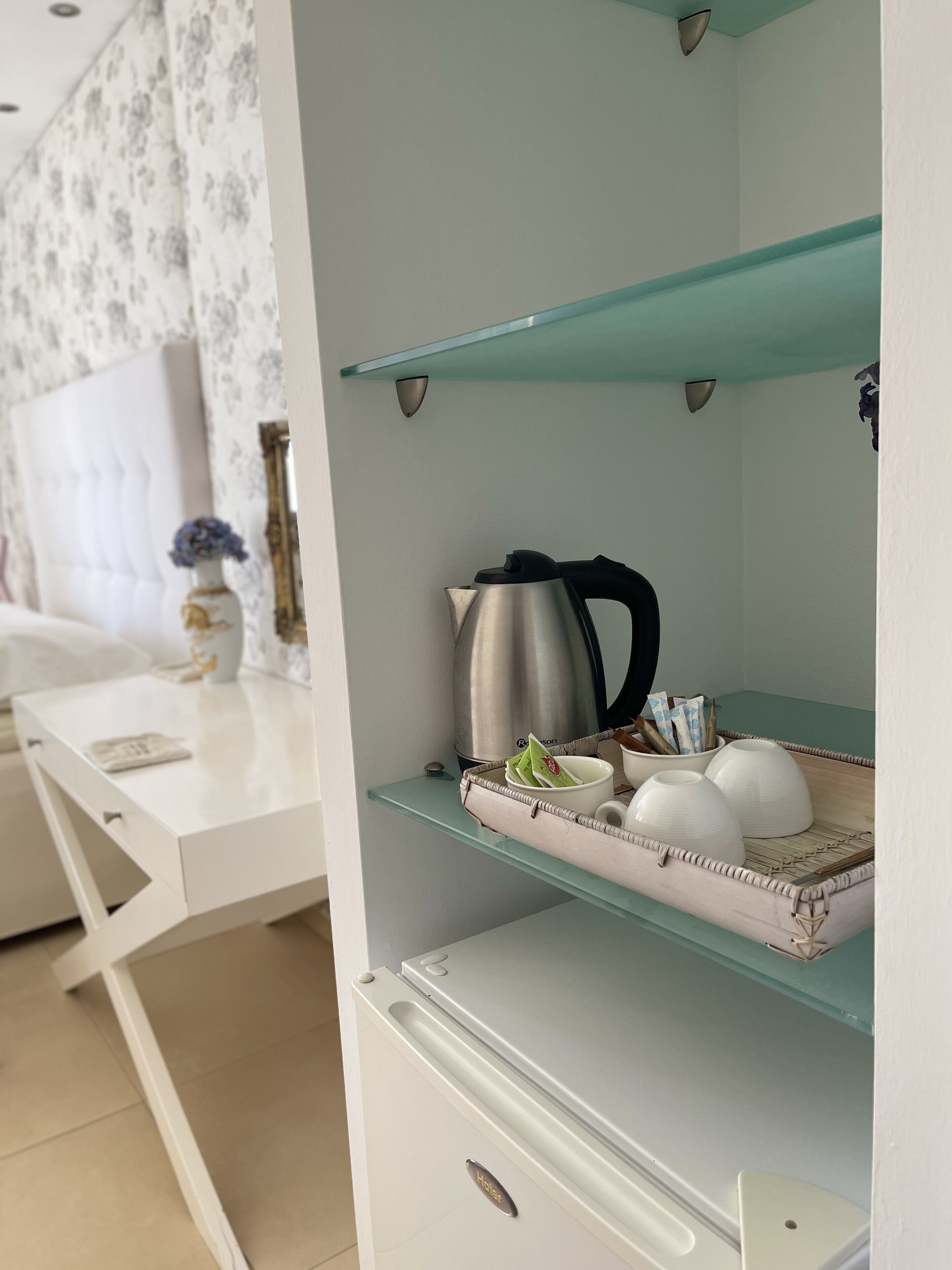 Et moderne hotelværelse med et hvidt skrivebord, en glashylde med elkedel og tesæt, samt en seng med blomstret sengegavl.