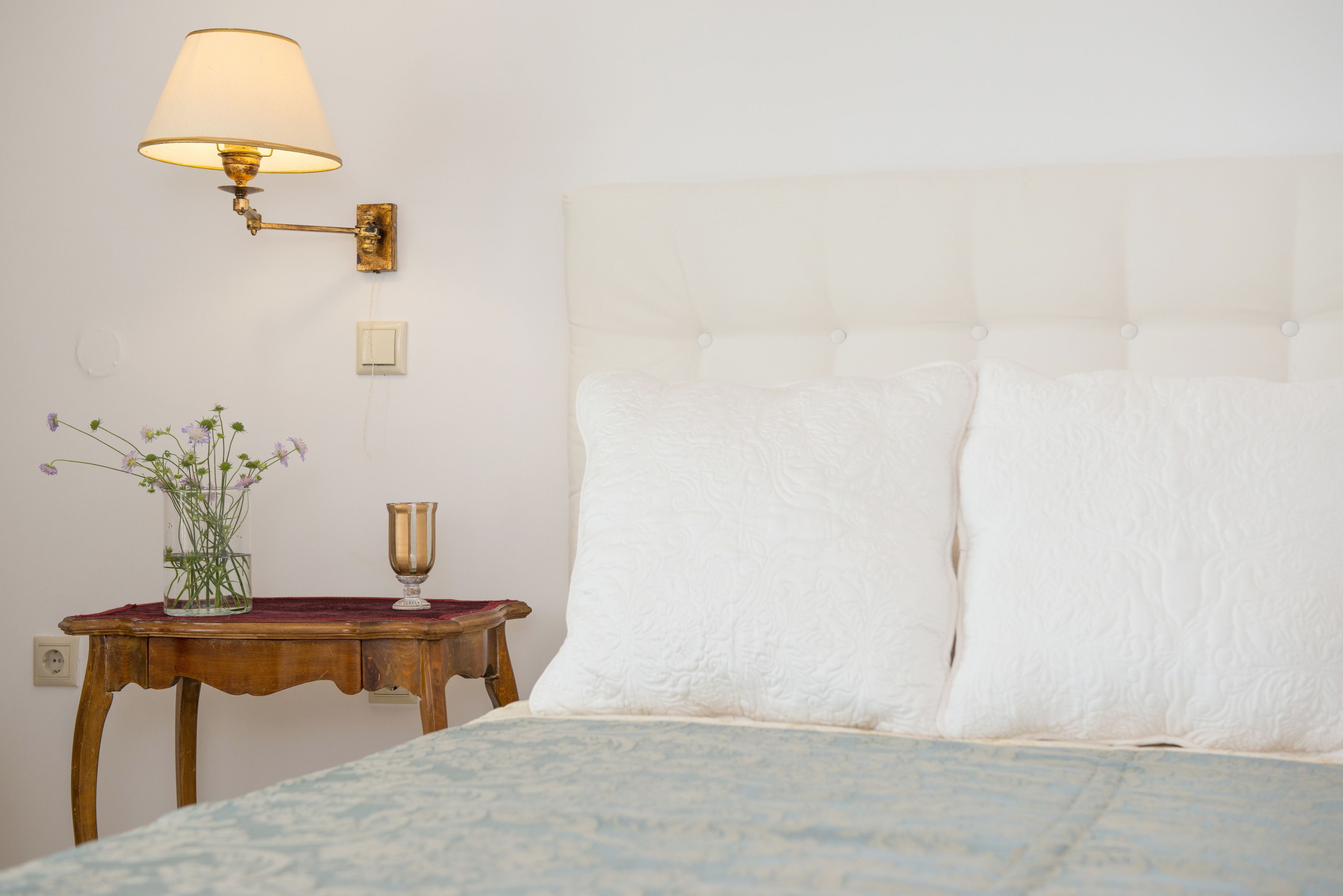 Un lit bien fait, avec une tête de lit blanche, une table de chevet sur laquelle se trouve une lampe, et un vase rempli de fleurs.