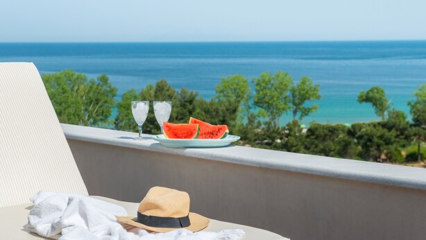 Un balcón con vista al mar, un plato con tajadas de sandía, dos vasos, un sombrero y una toalla.