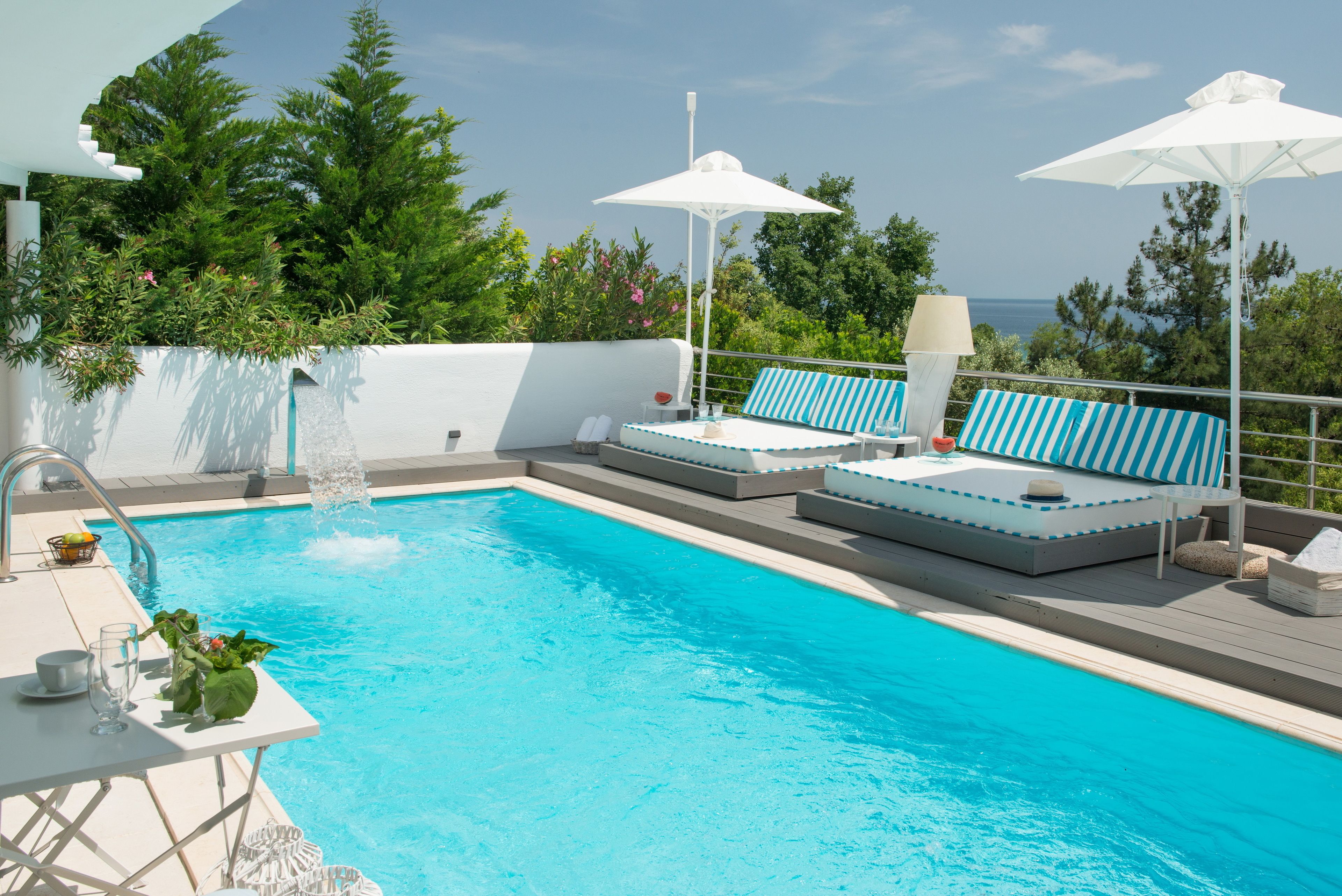 Piscina con sillas de descanso y sombrillas, rodeada de follaje y con vistas a una masa de agua.