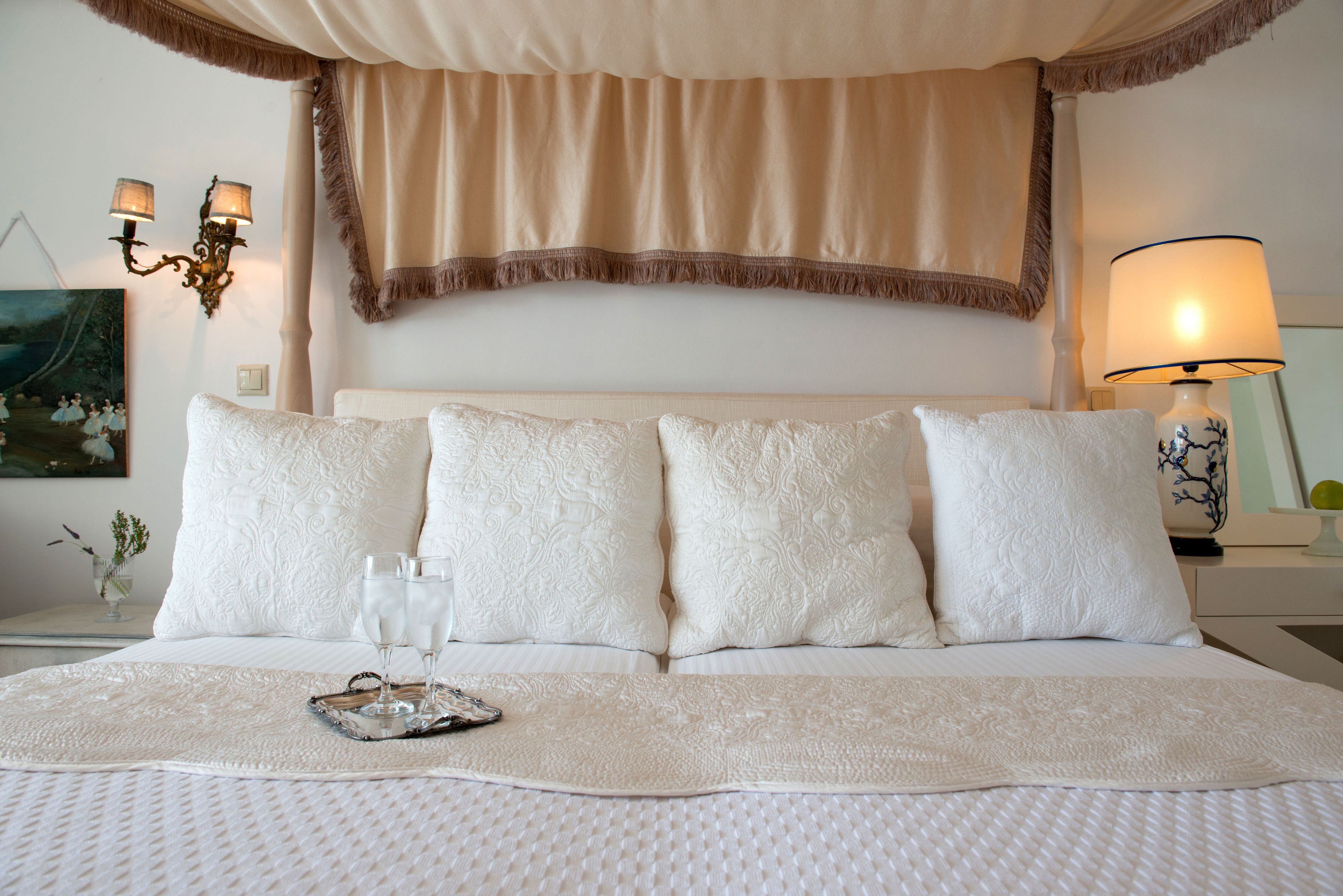 Cama arrumada com edredom branco, quatro travesseiros e bandeja com copos e controle remoto.