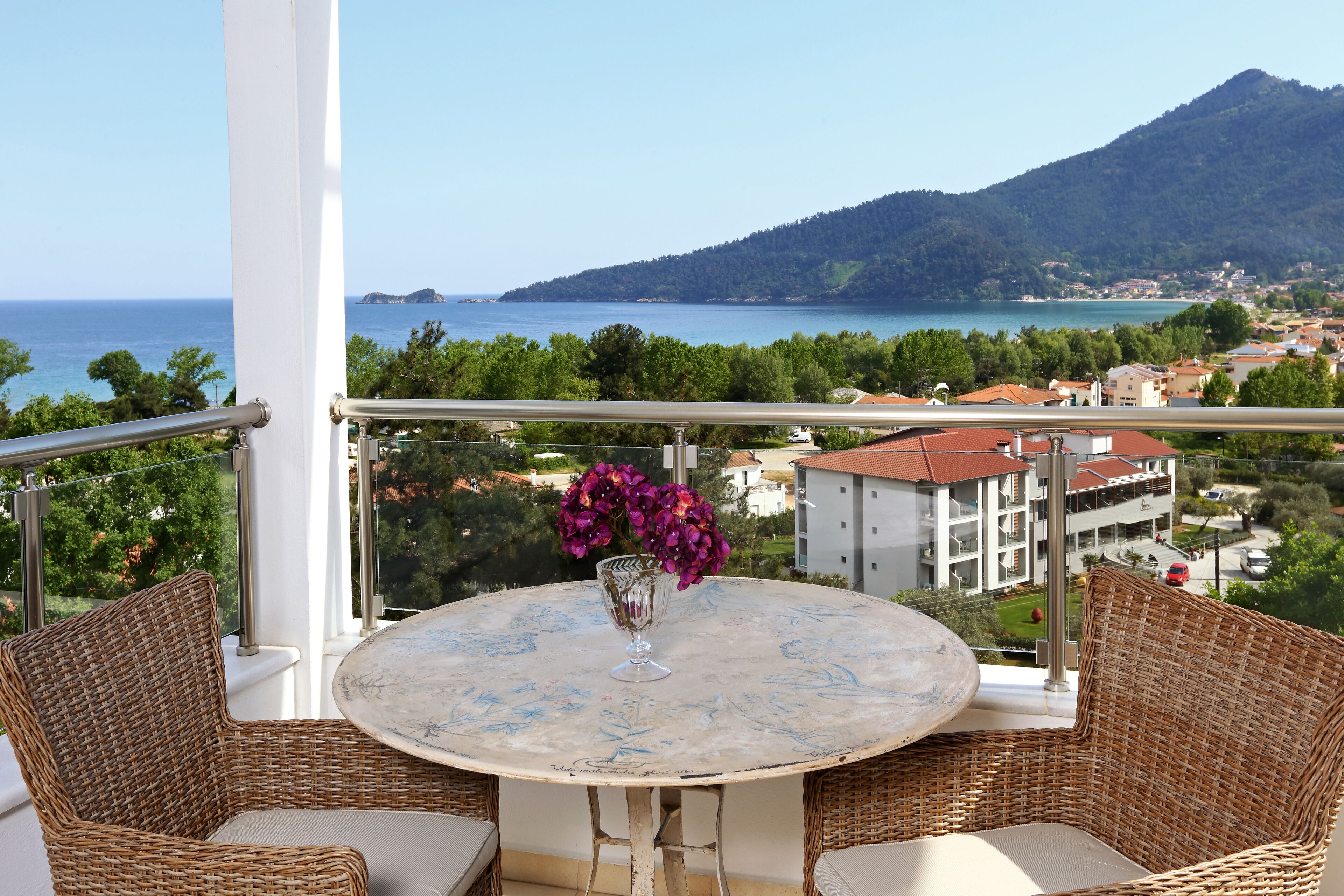 Un balcón con mobiliario de mimbre, una mesa con un florero y vistas al mar y a las montañas.