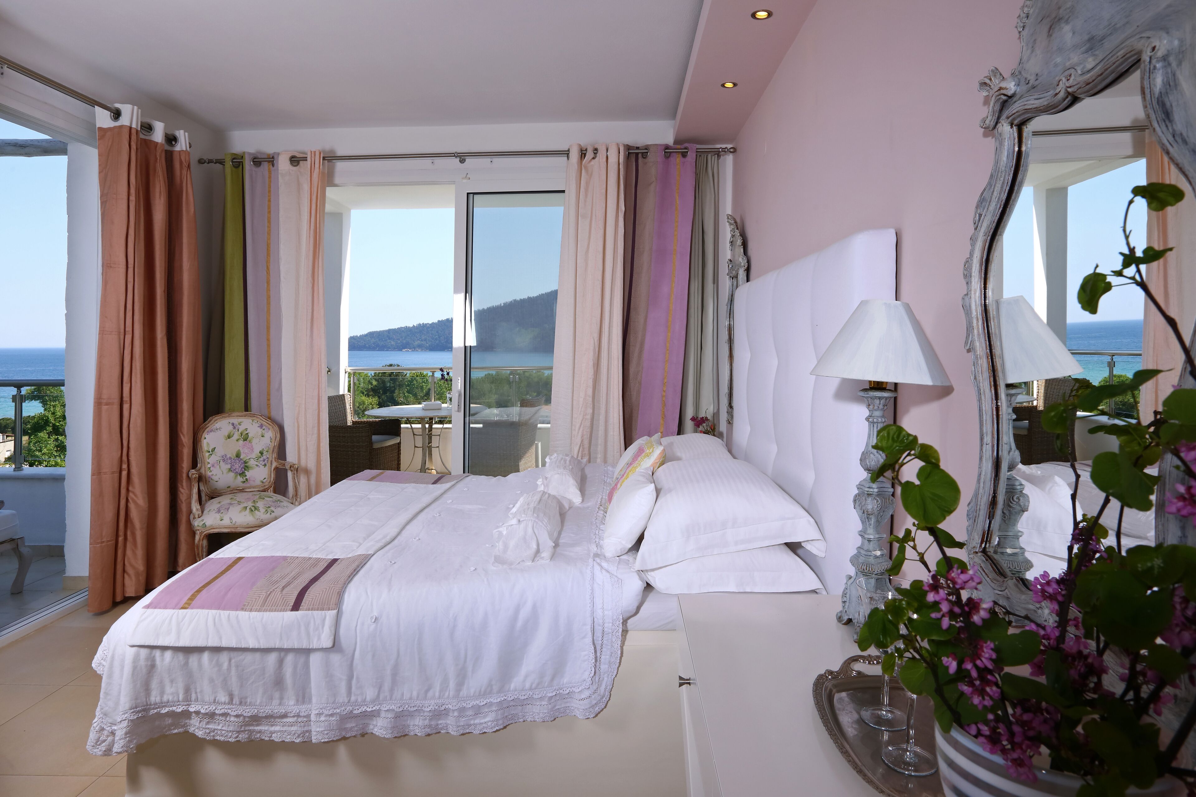 A bedroom with a large bed, a chair, a table, a mirror, and a view of the sea.