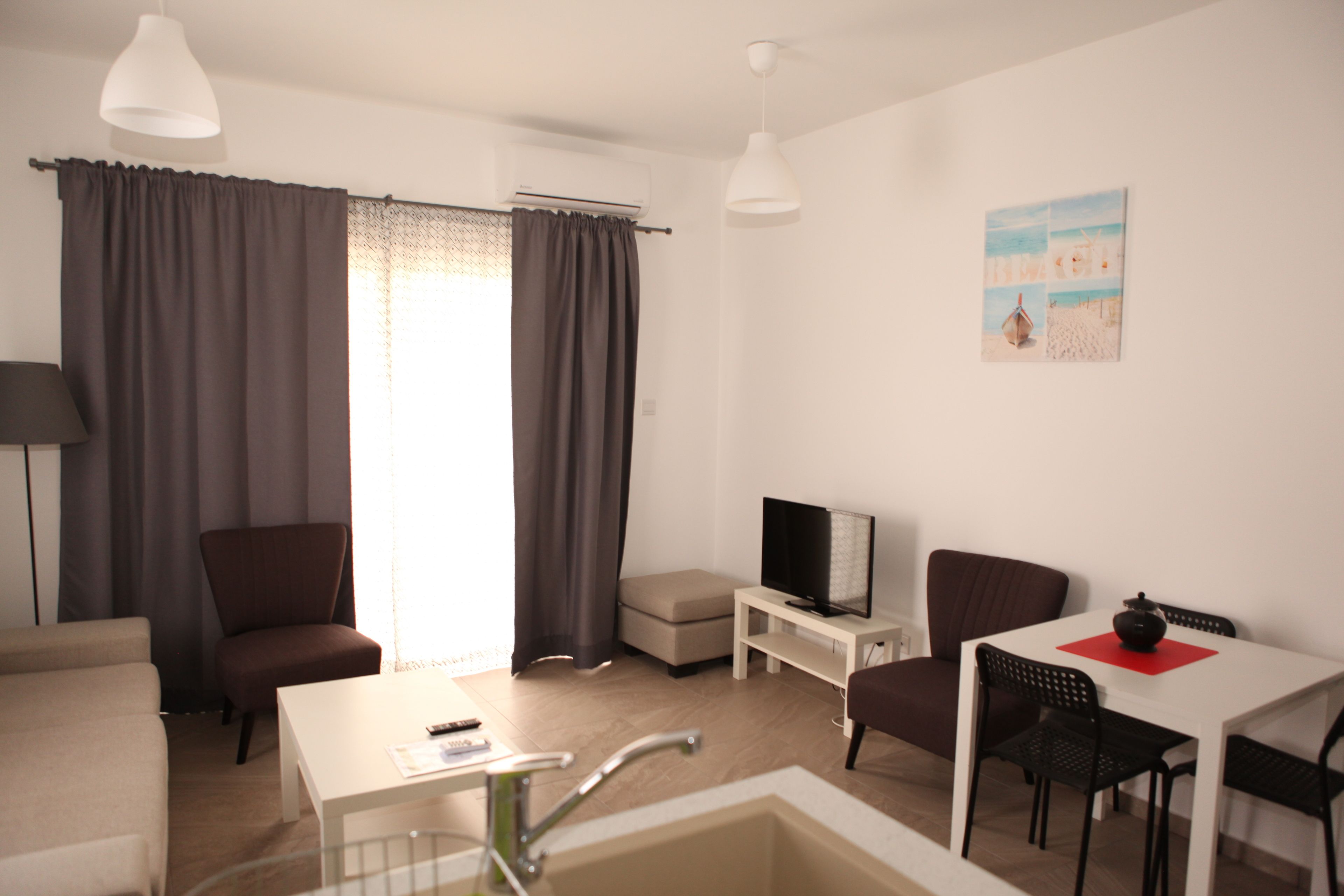 Una habitación de hotel moderna con sofá, televisión, mesa de comedor con sillas, una mesita con control remoto y un cuadro en la pared.