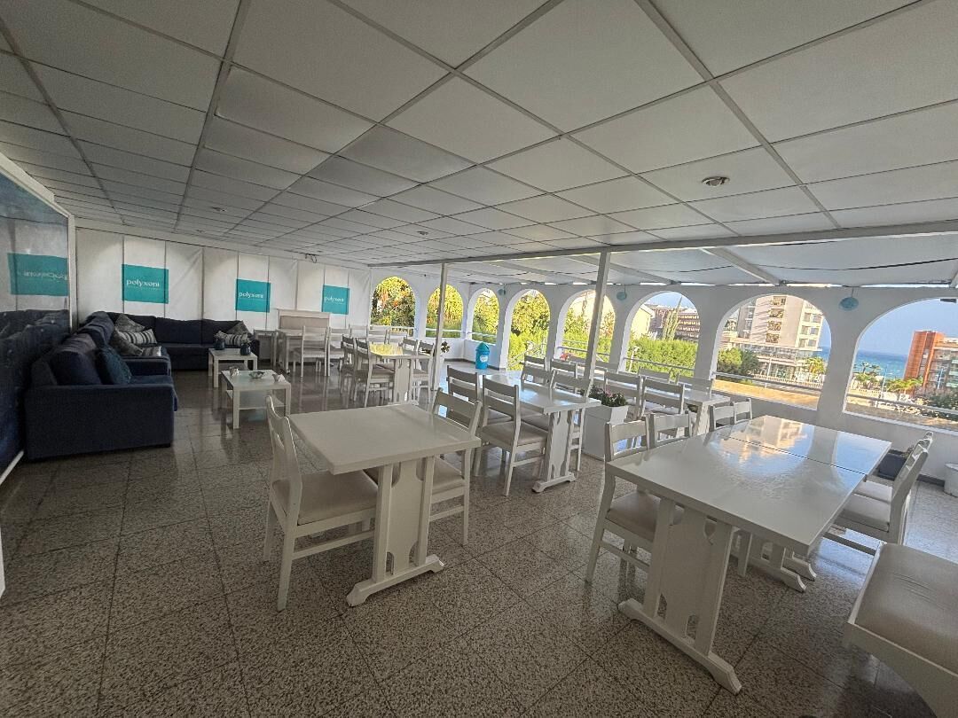 Un amplio espacio interior con varias mesas y sillas blancas, un sofá y vistas a través de grandes ventanales.