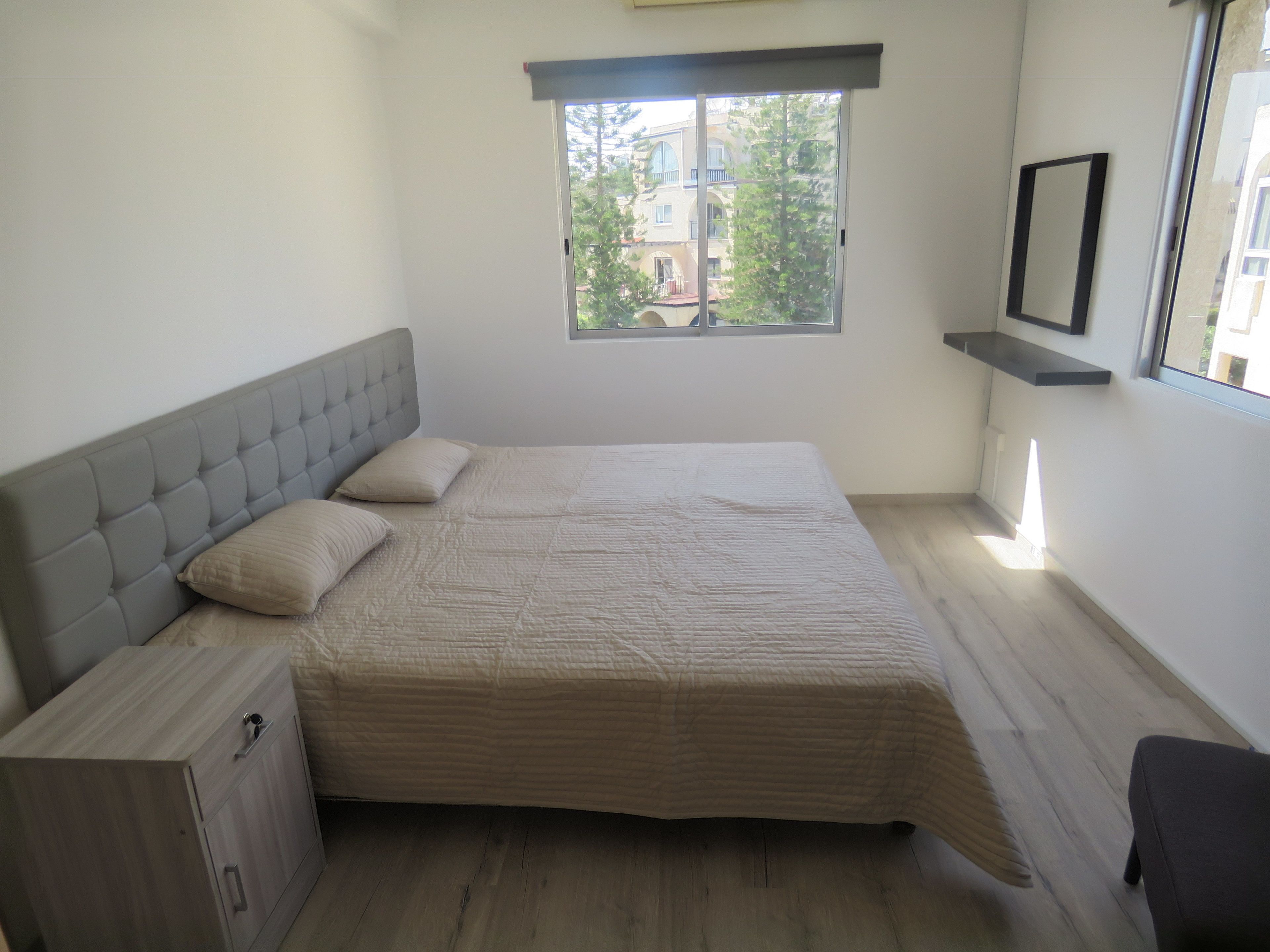 Ein Schlafzimmer mit einem großen Bett, einem Nachttisch, einem Kleiderschrank, einem Fenster mit Blick auf Gebäude und einem Spiegelregal.