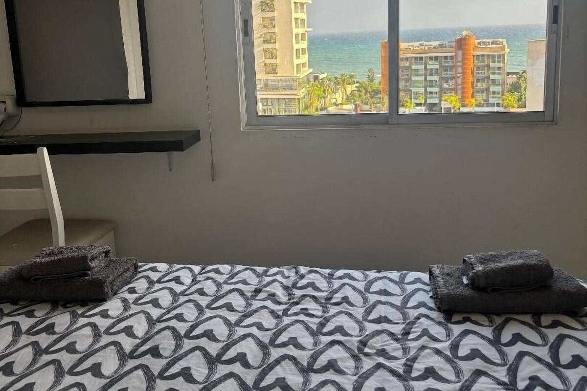 A hotel room with a bed, a desk, and a large window offering a view of buildings and the sea.