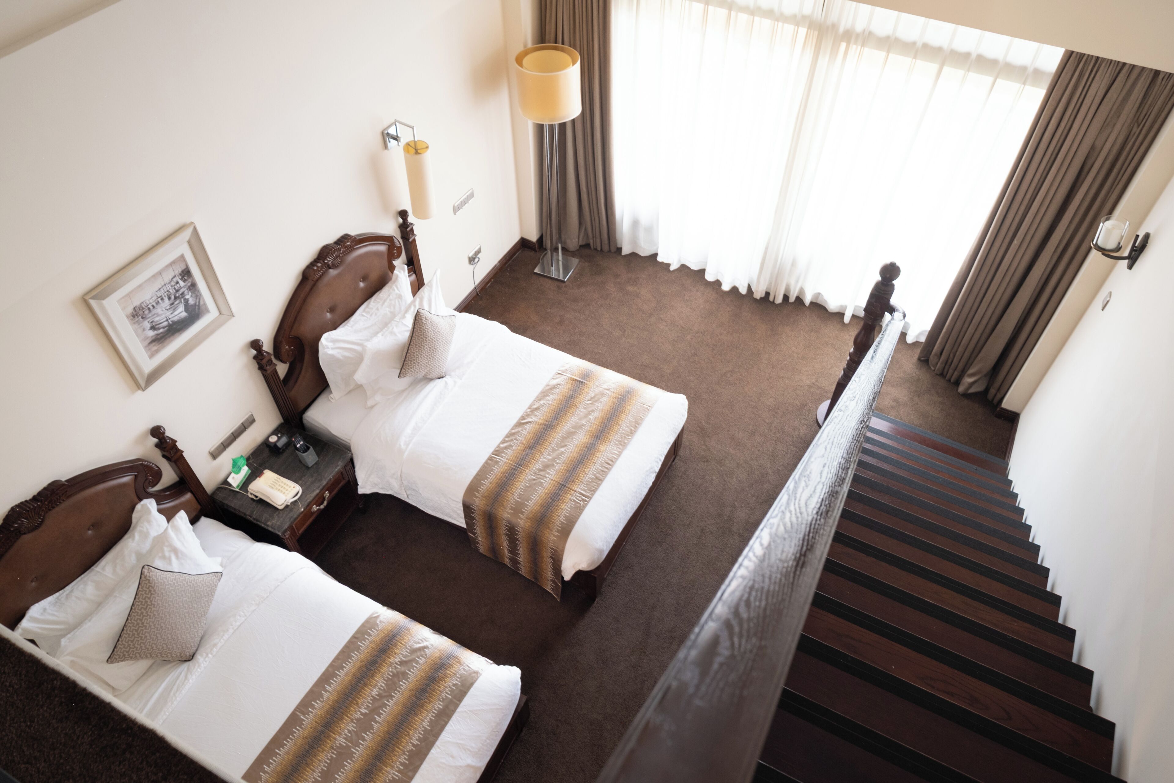 A hotel room with two beds, a nightstand, a lamp, a framed picture on the wall, and a staircase.