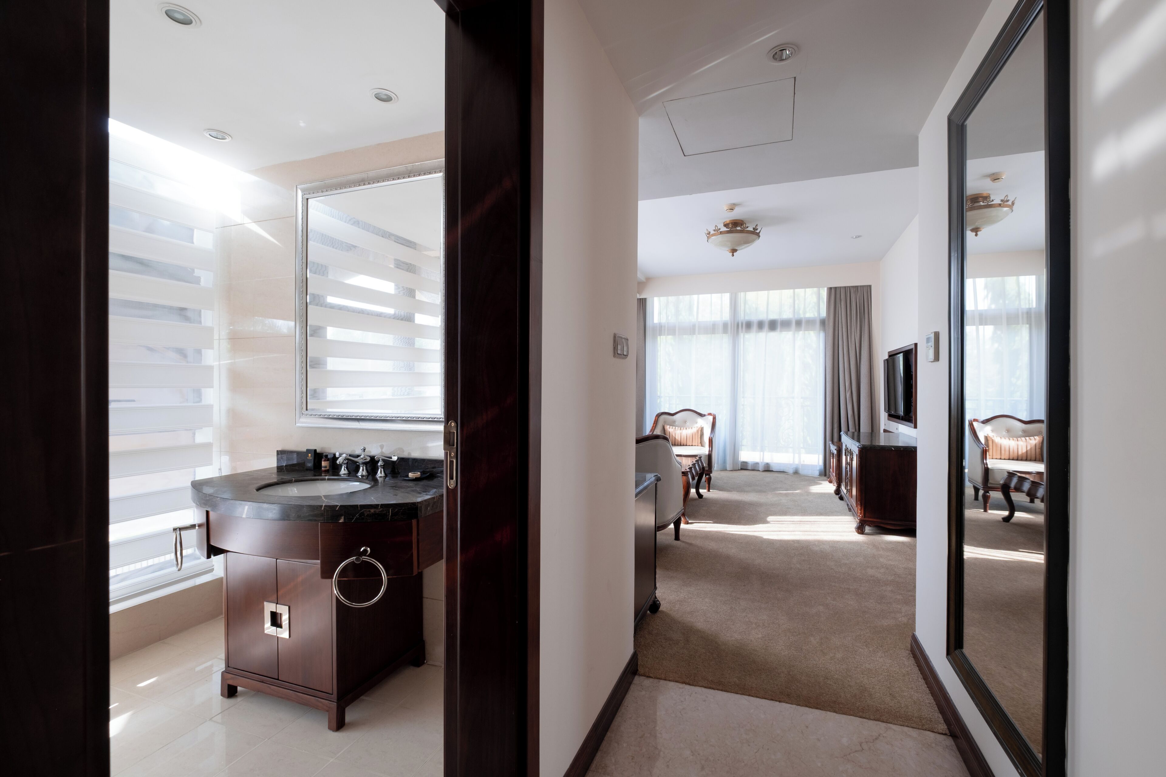 A modern hotel room with a large mirror, a dark wood vanity, a large window with blinds, a sofa, and a television.