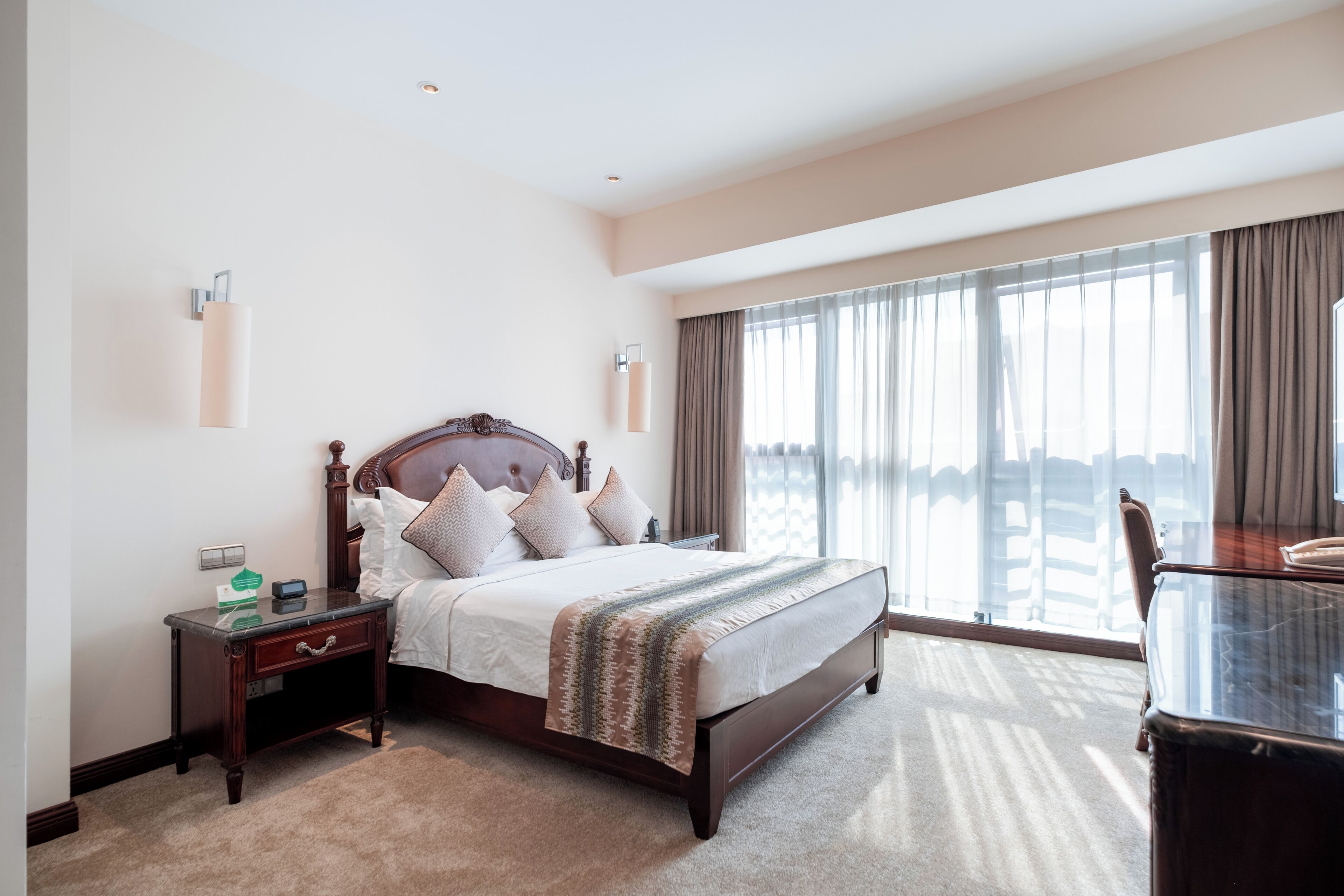 Quarto de hotel com uma cama grande, uma mesa de cabeceira, uma escrivaninha e vista da cidade através de amplas janelas.