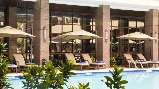 Zona de piscina con sillones de descanso y sombrillas, rodeada por un edificio con amplios ventanales y pilares de piedra.