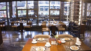 Un restaurante con varias mesas y sillas, ventanas amplias que ofrecen vistas de la ciudad, y un diseño de interiores moderno.