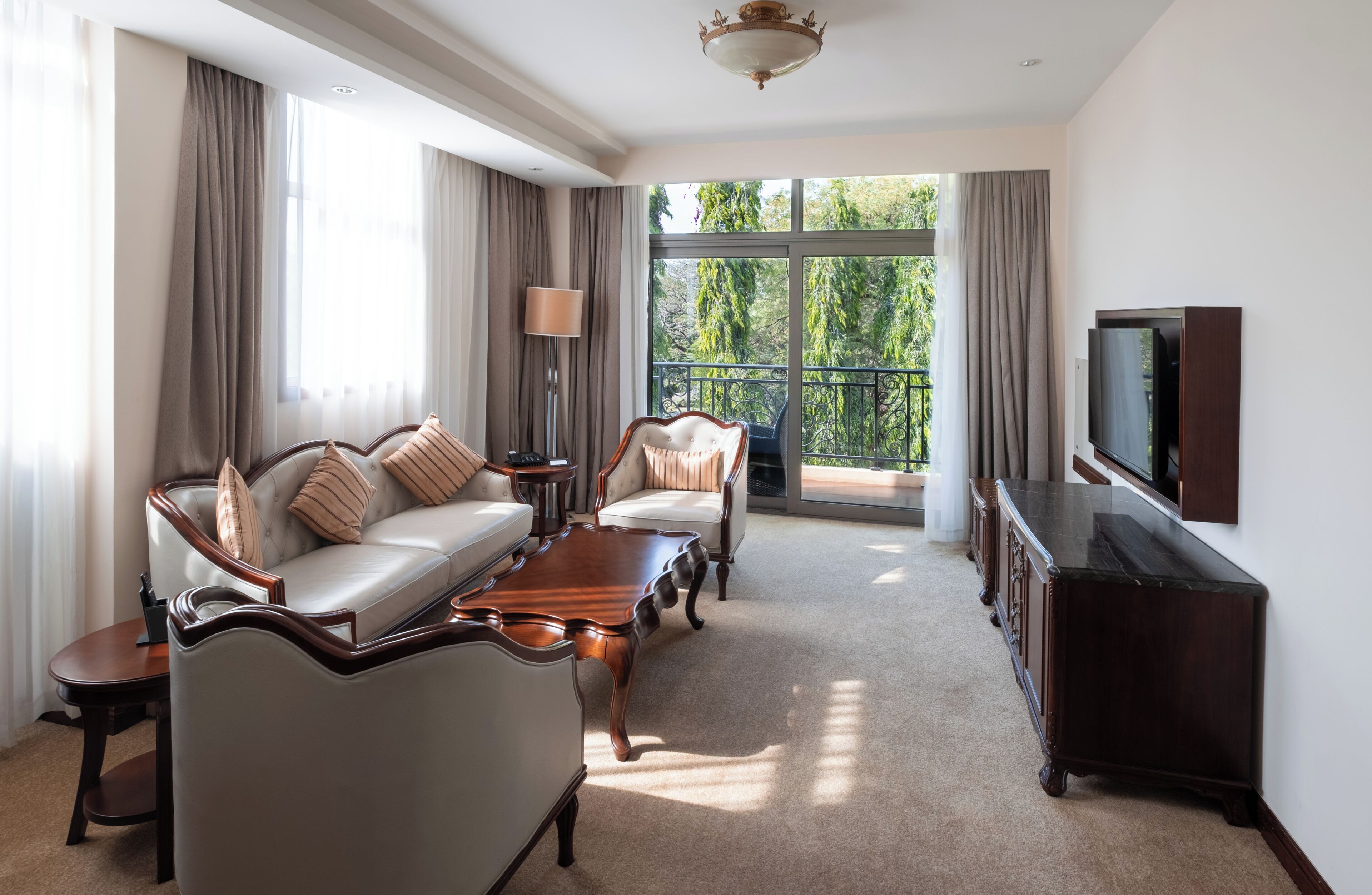 A spacious living room with a sofa, armchairs, a coffee table, a TV, and a balcony with a view of trees.