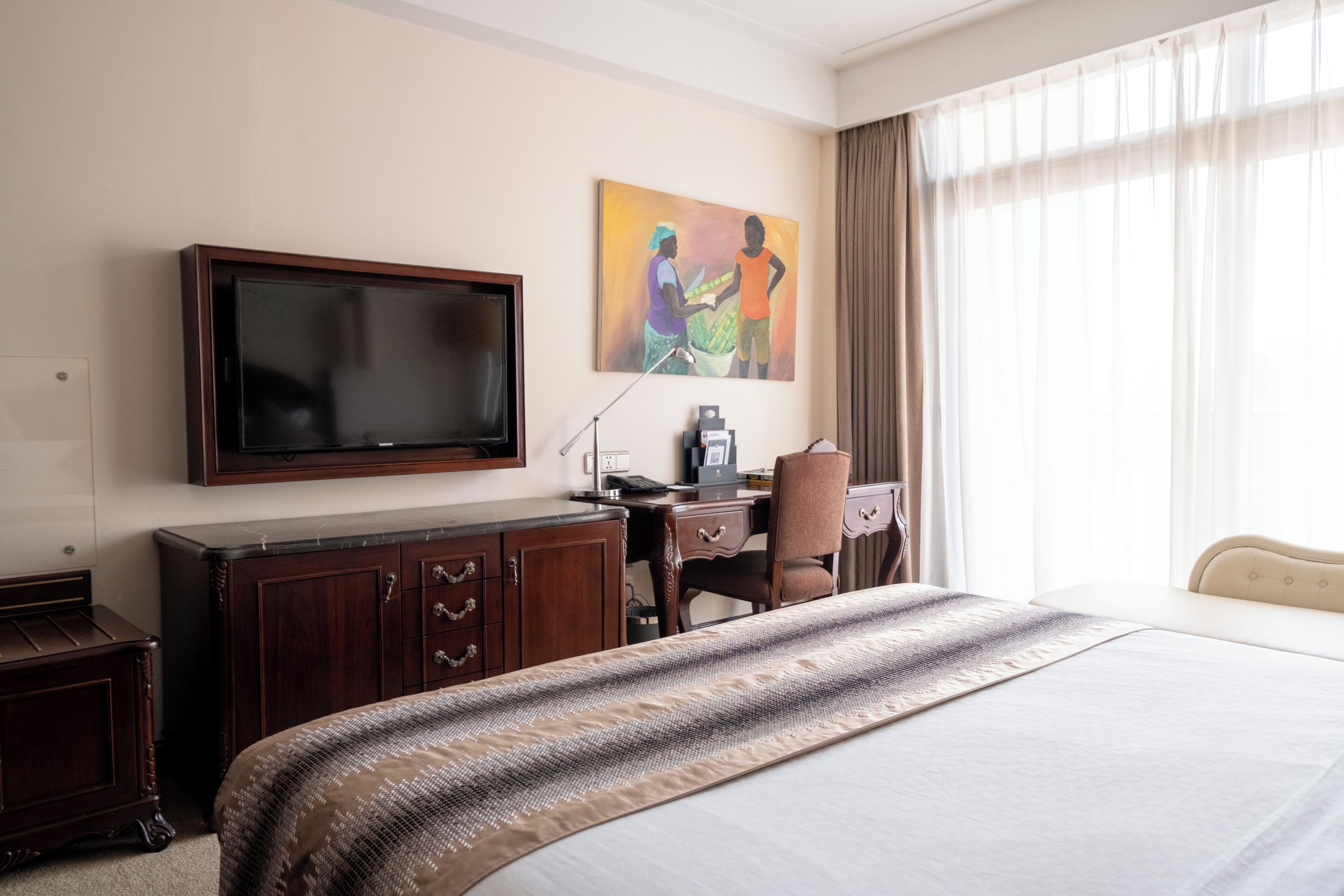 A hotel room with a large bed, a desk with a chair, a television mounted on the wall, and a painting above the desk.
