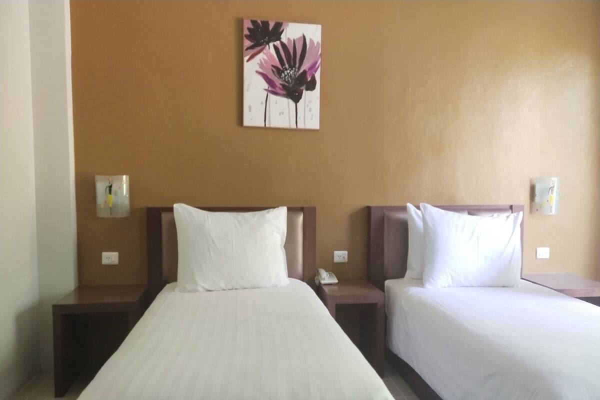 Habitación de hotel con dos camas individuales, cada una con cabecero de madera y ropa de cama blanca. Hay una obra de arte colgada en la pared sobre las camas y una mesita de noche con un teléfono a cada lado.