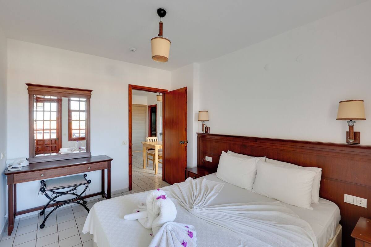 Habitación de hotel con cama, escritorio, silla, espejo y puerta que da a un comedor.