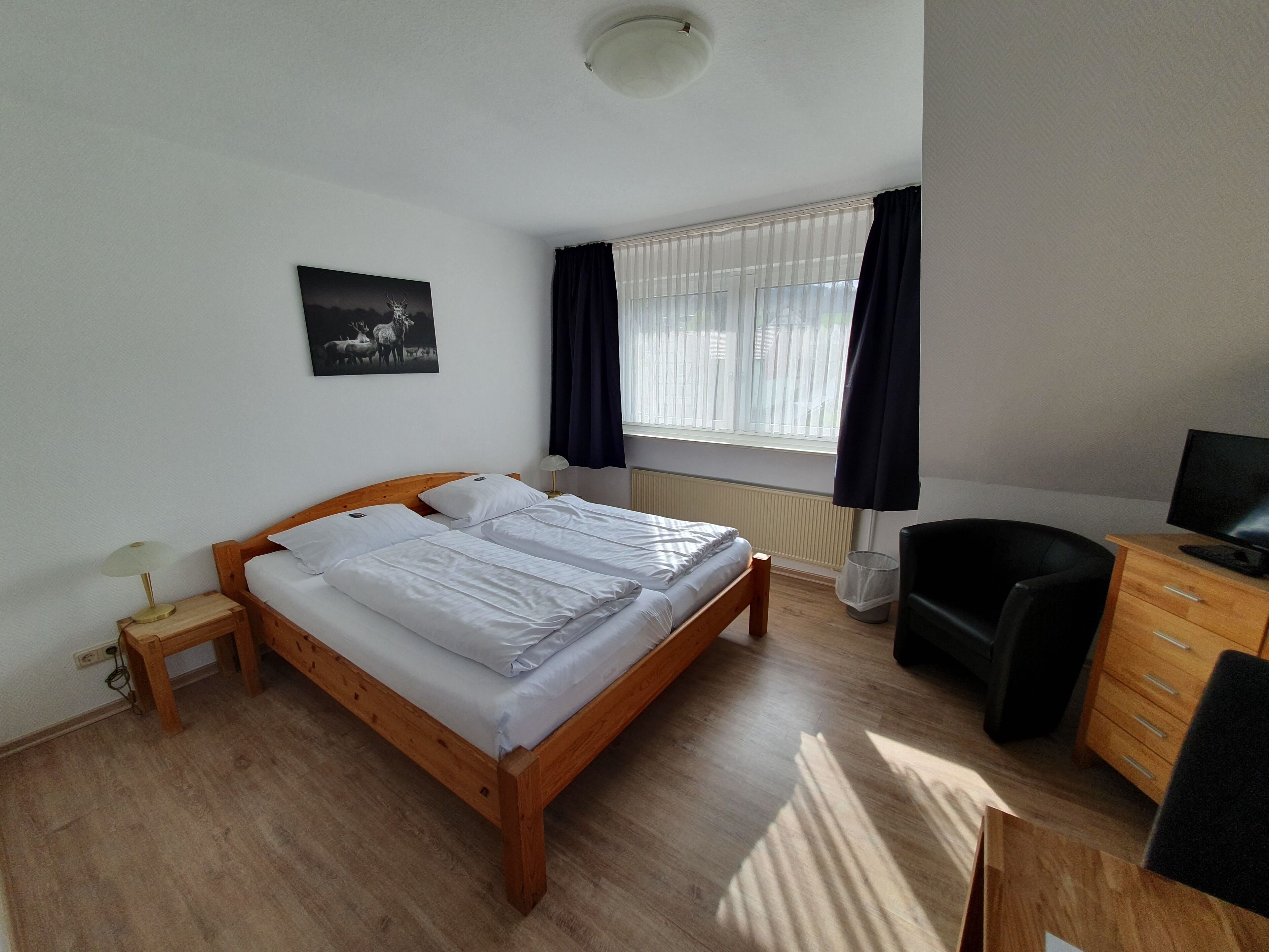 A bedroom with a wooden bed, a desk with a chair, a window with curtains, a radiator, and a wall-mounted picture.
