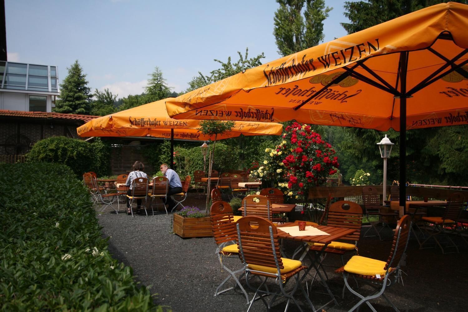 An outdoor seating area with tables and chairs under orange umbrellas, a flower arrangement, and a building in the background.