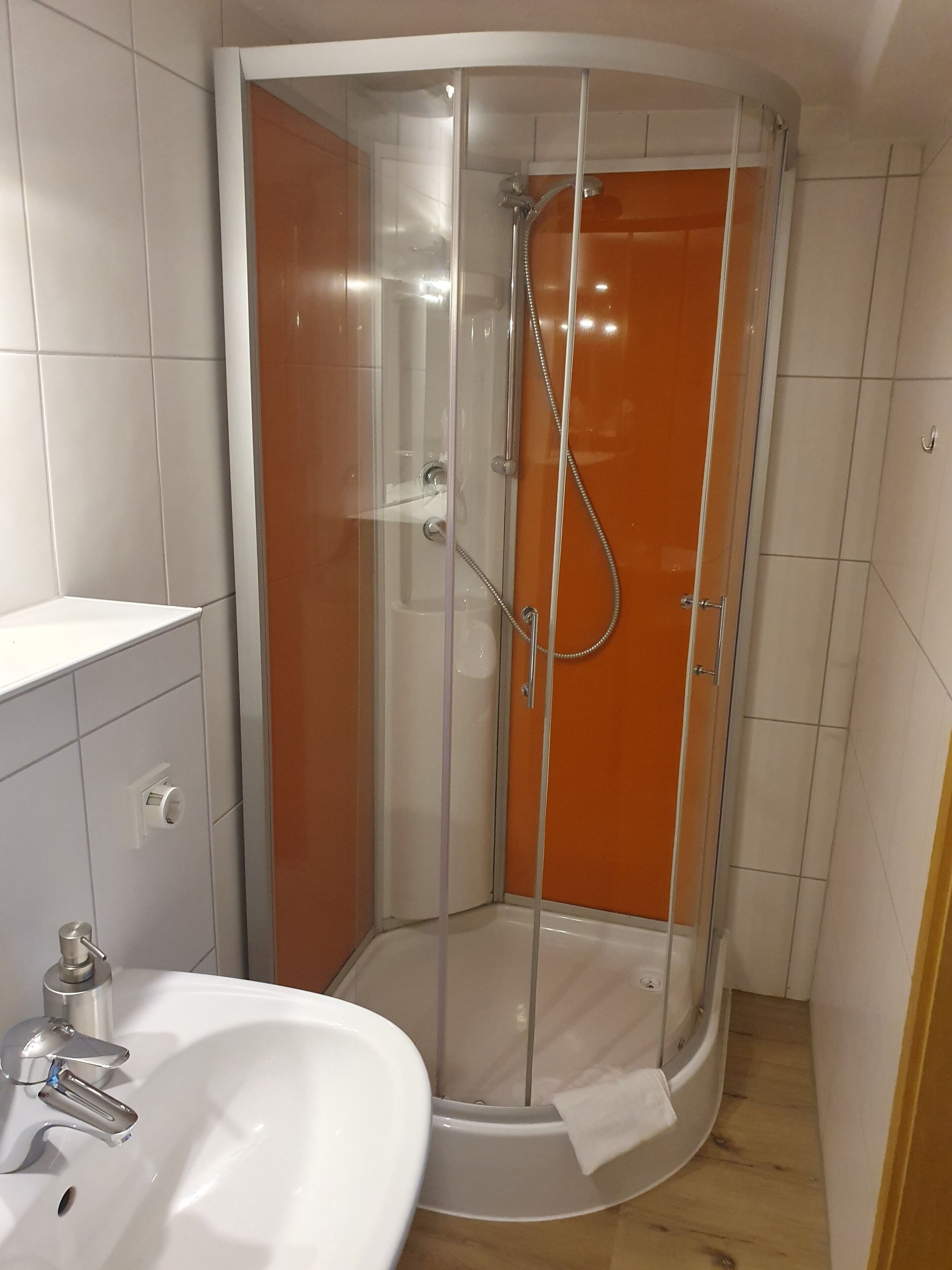 A corner bathroom with a glass-enclosed shower, a white sink, and a wooden floor.