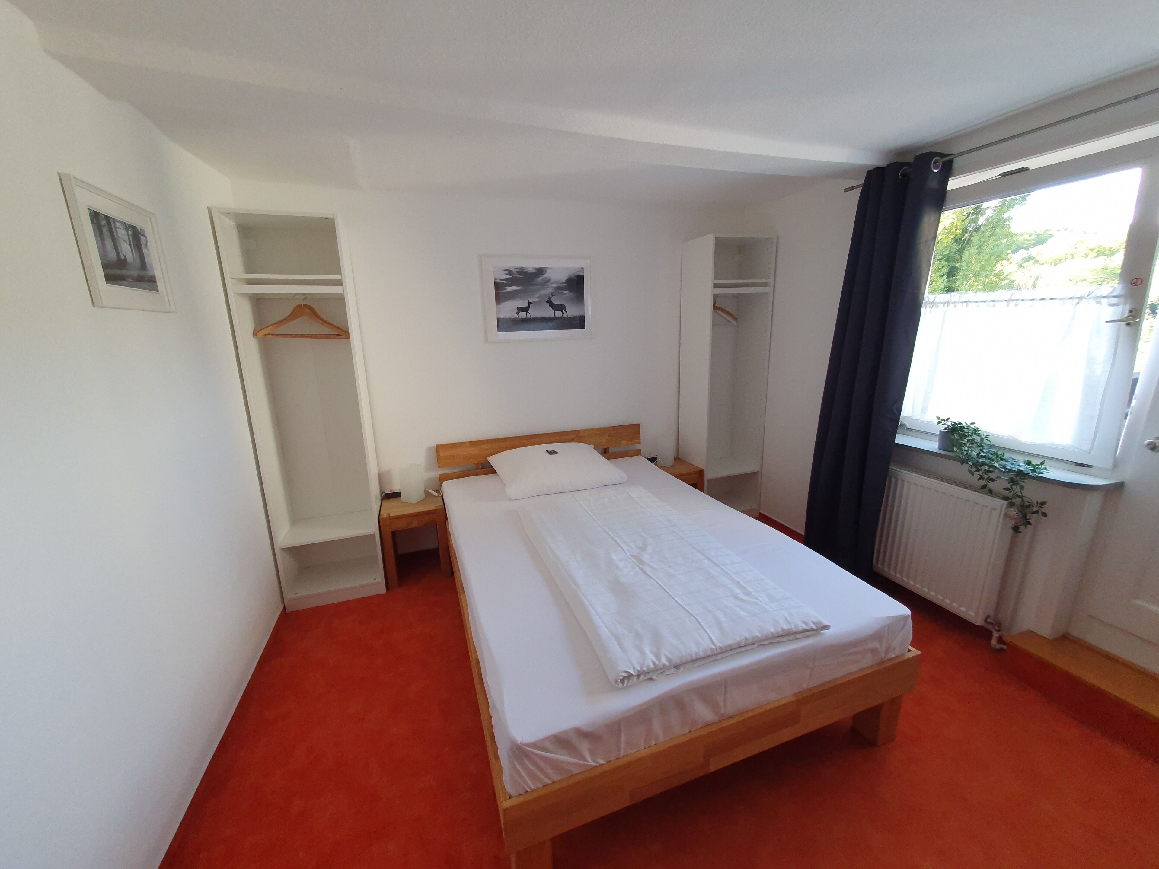 A neatly arranged bedroom with a single bed, a bedside table, a hanging wardrobe, and a window with a view of greenery.