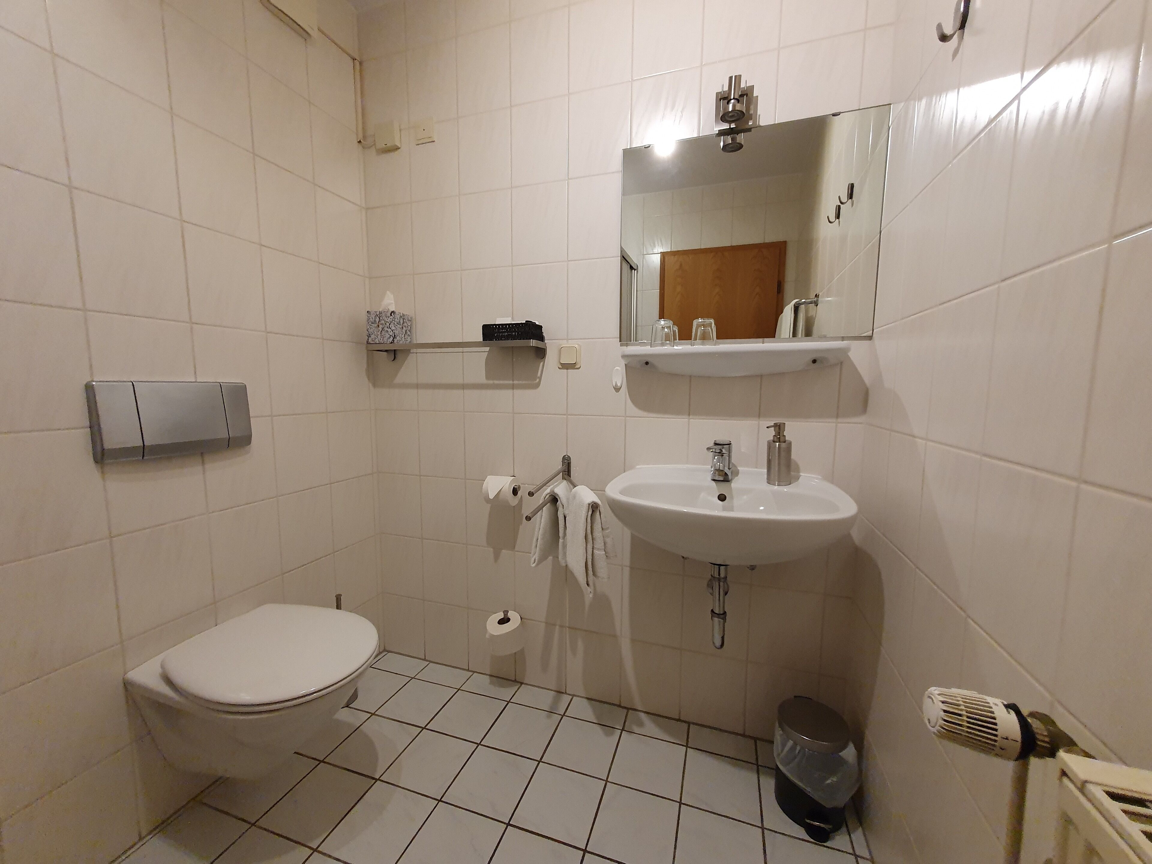 Un baño pequeño y limpio, con inodoro, lavabo y espejo. Hay repisas sobre el espejo con artículos de aseo.