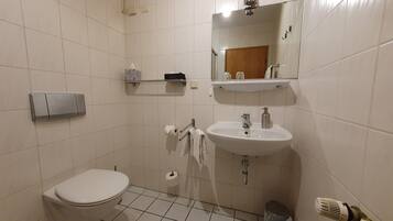 Un baño pequeño y limpio, con inodoro, lavabo y espejo. Hay repisas sobre el espejo con artículos de aseo.