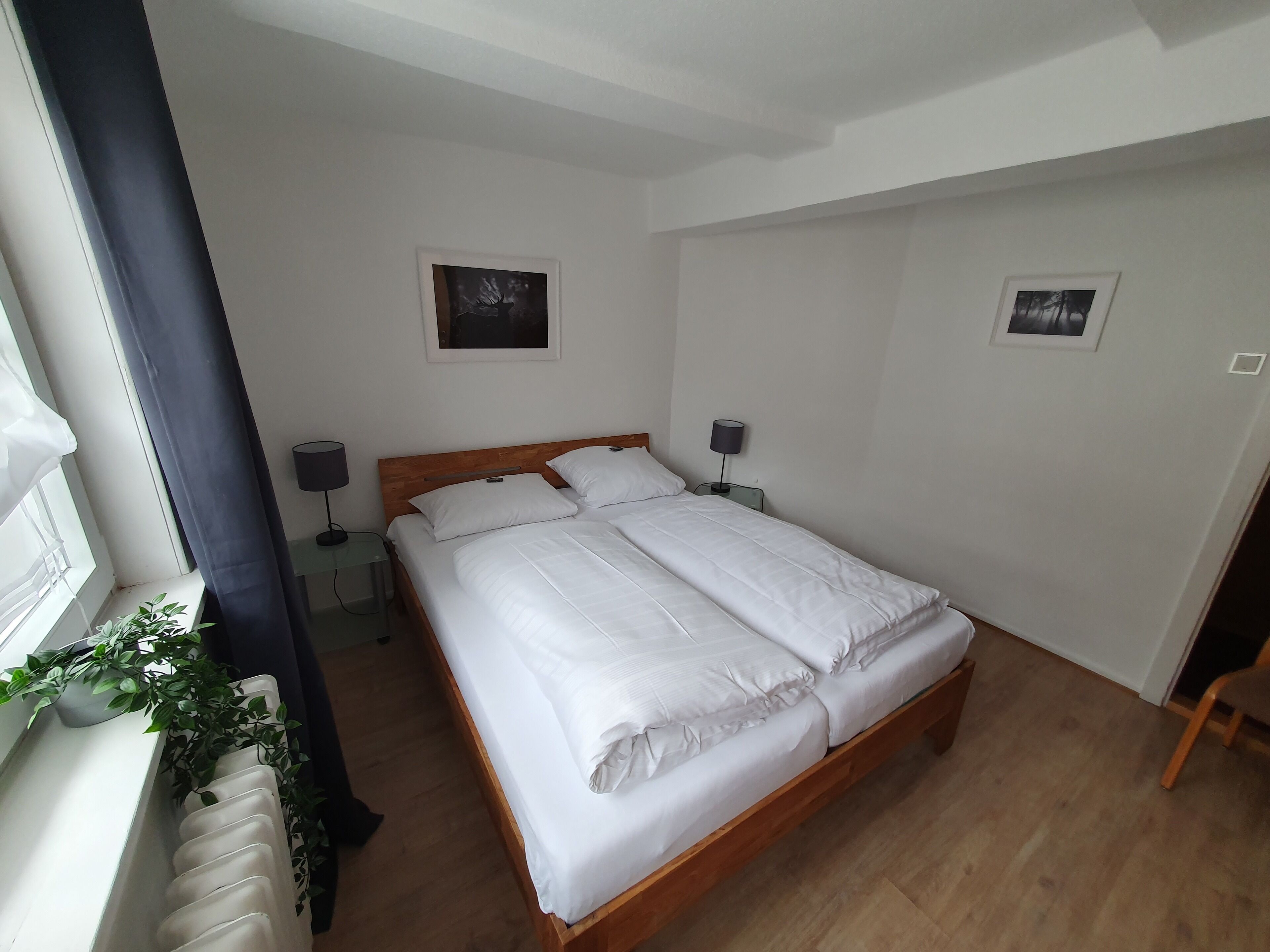 Un llit ben arrenjat amb llençols blancs en una habitació amb terres de fusta, una finestra amb cortina i dos quadres penjats a la paret.