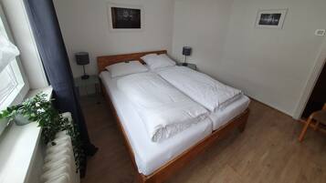 Un llit ben arrenjat amb llençols blancs en una habitació amb terres de fusta, una finestra amb cortina i dos quadres penjats a la paret.