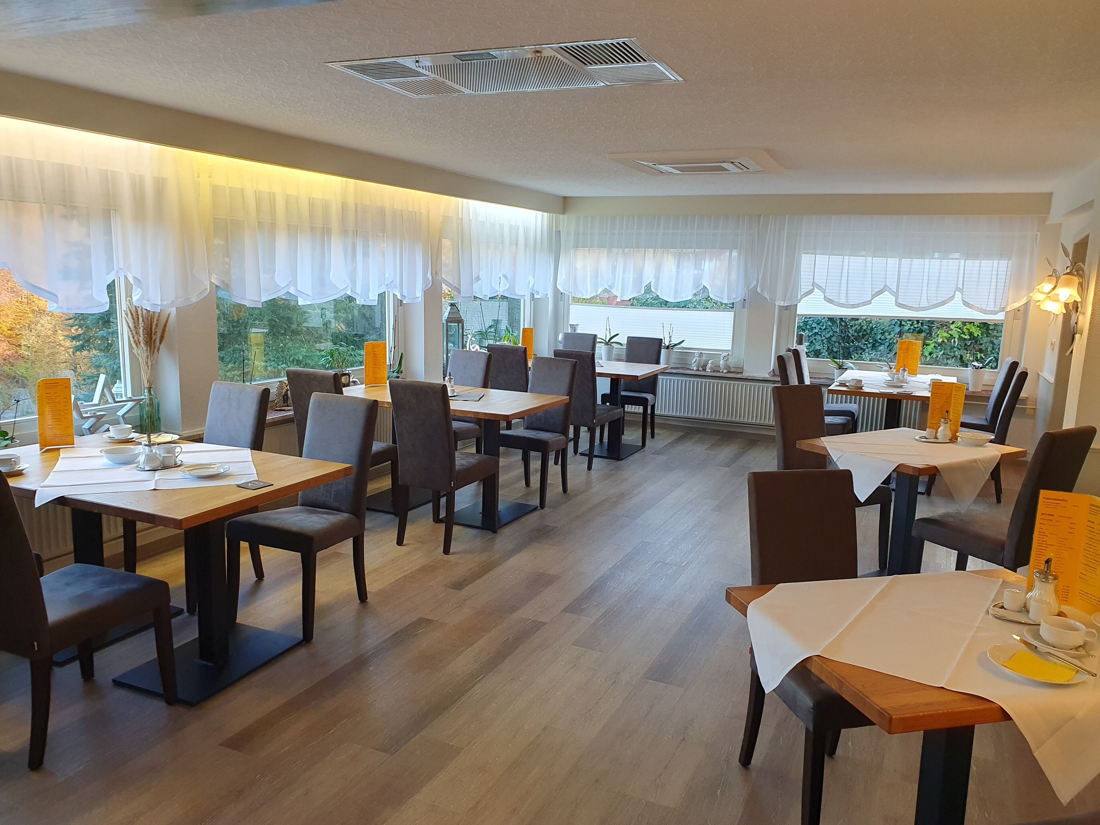 A restaurant with multiple tables and chairs, large windows with sheer curtains, and a view of greenery outside.