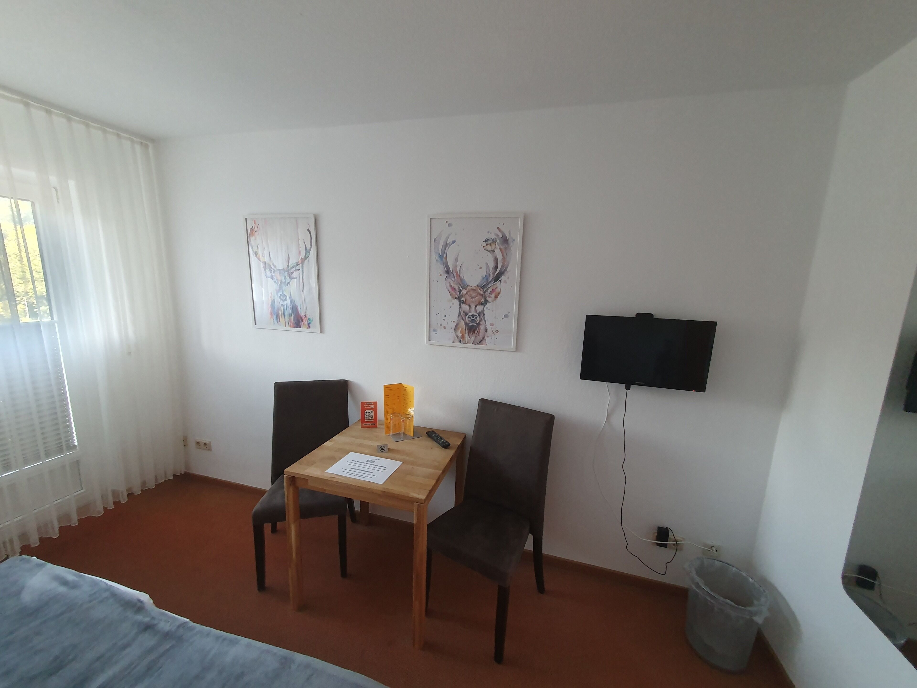 Habitación de hotel con una cama, una mesa con una silla, un televisor montado en la pared y dos cuadros enmarcados.